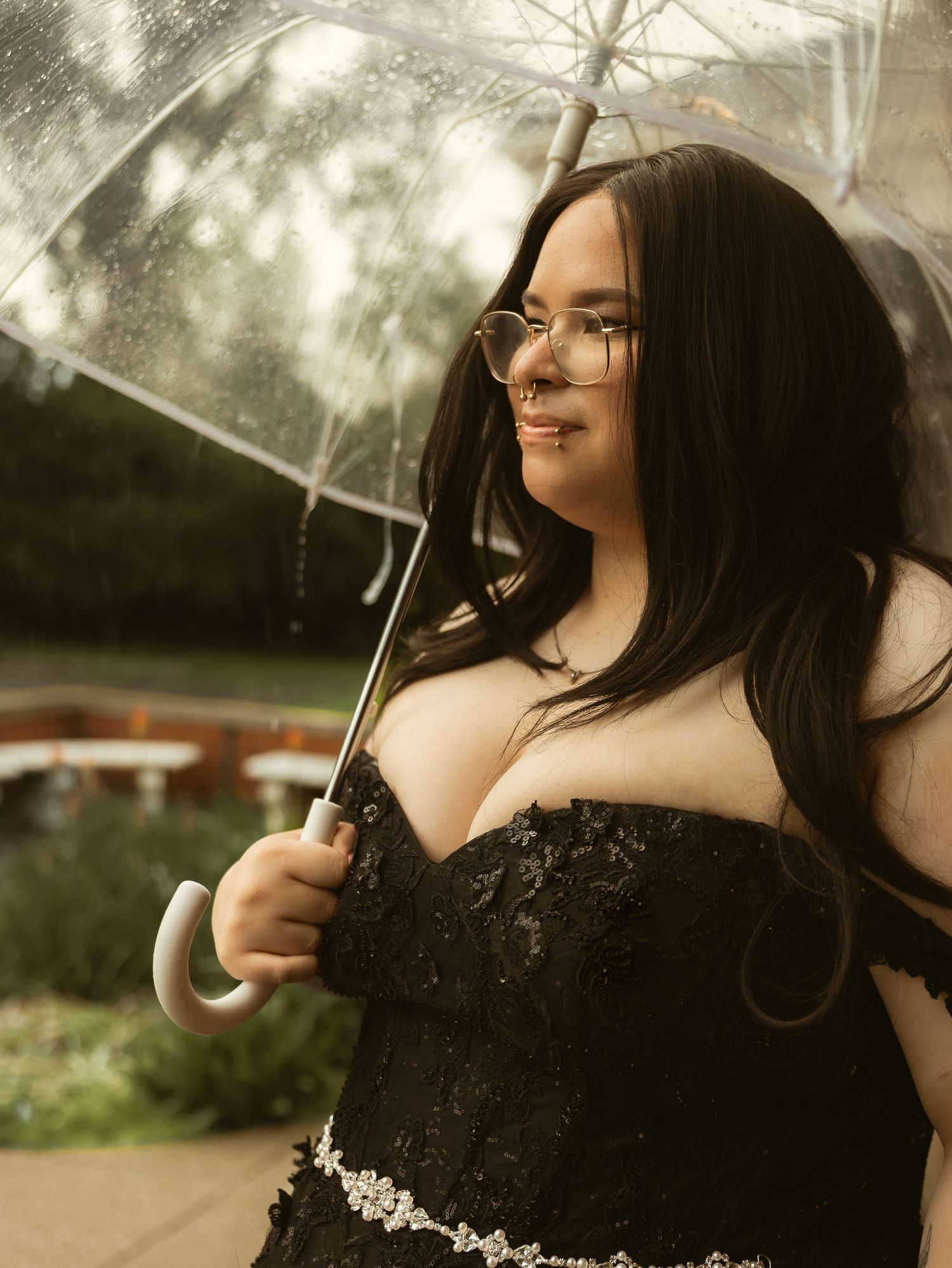 Bride in black lace corset wedding dress holds clear umbrella outdoors on rainy wedding day.