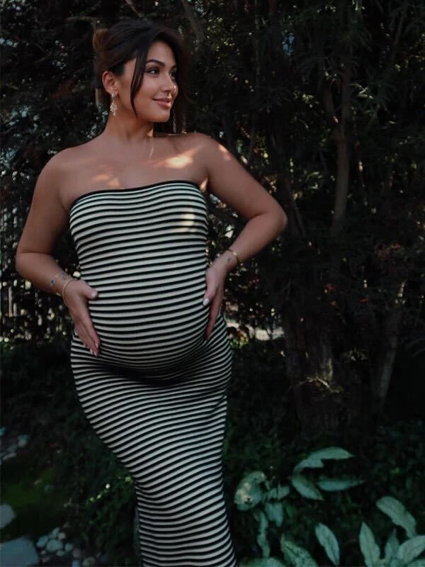A pregnant woman in a striped strapless dress poses outdoors against a dark foliage background.