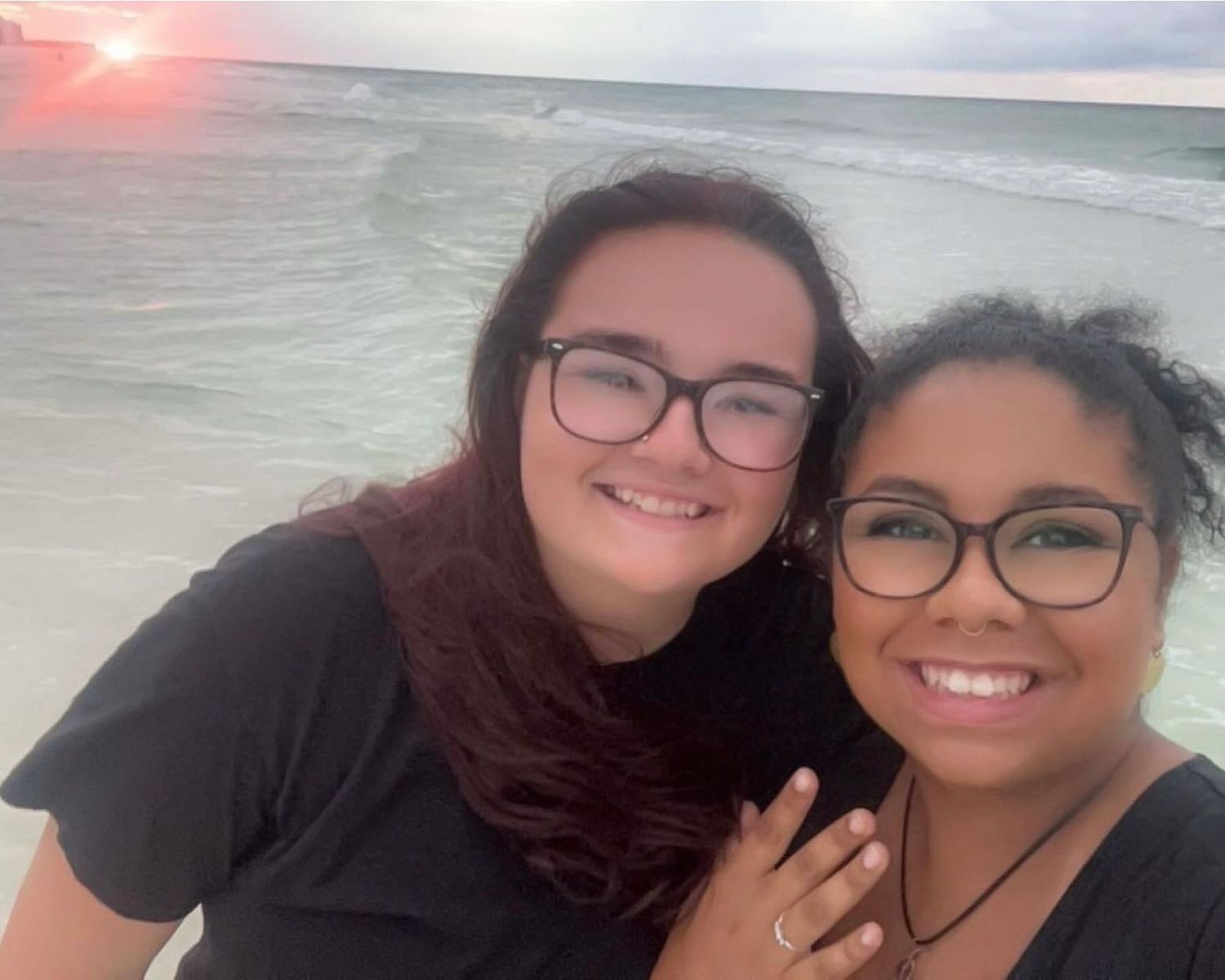 Two friends taking a beach selfie at sunset with ocean waves visible behind them.