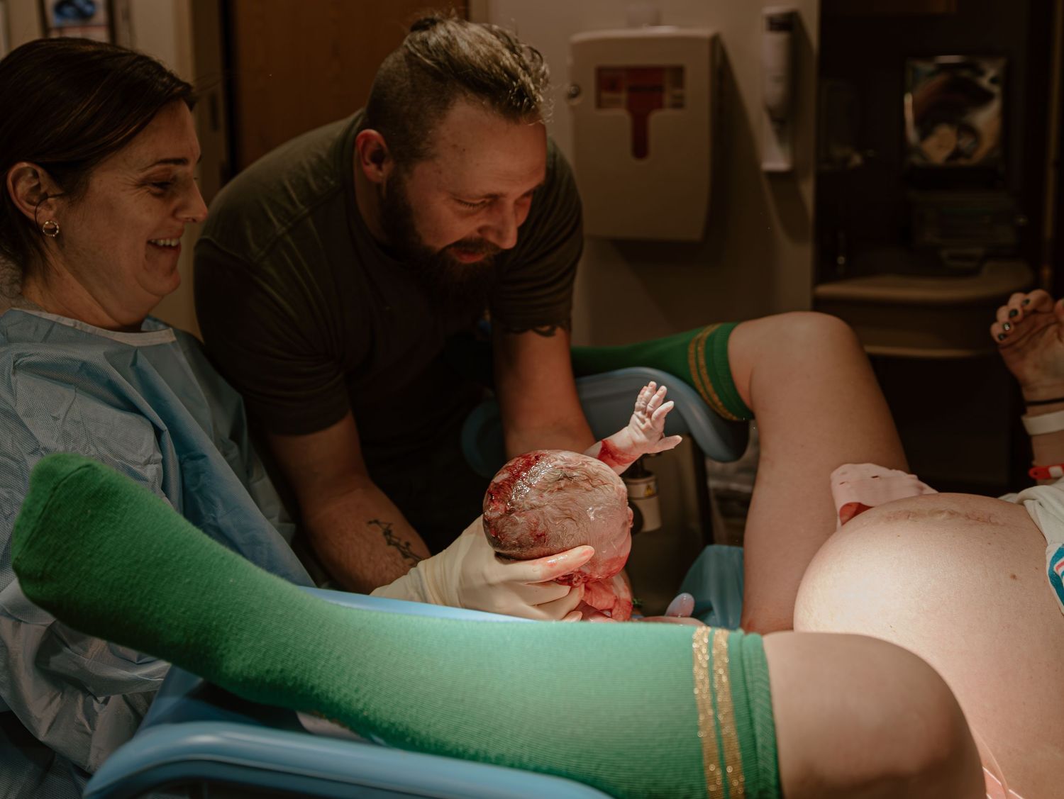 A newborn baby emerges during childbirth as medical professionals assist in a dimly lit delivery room.