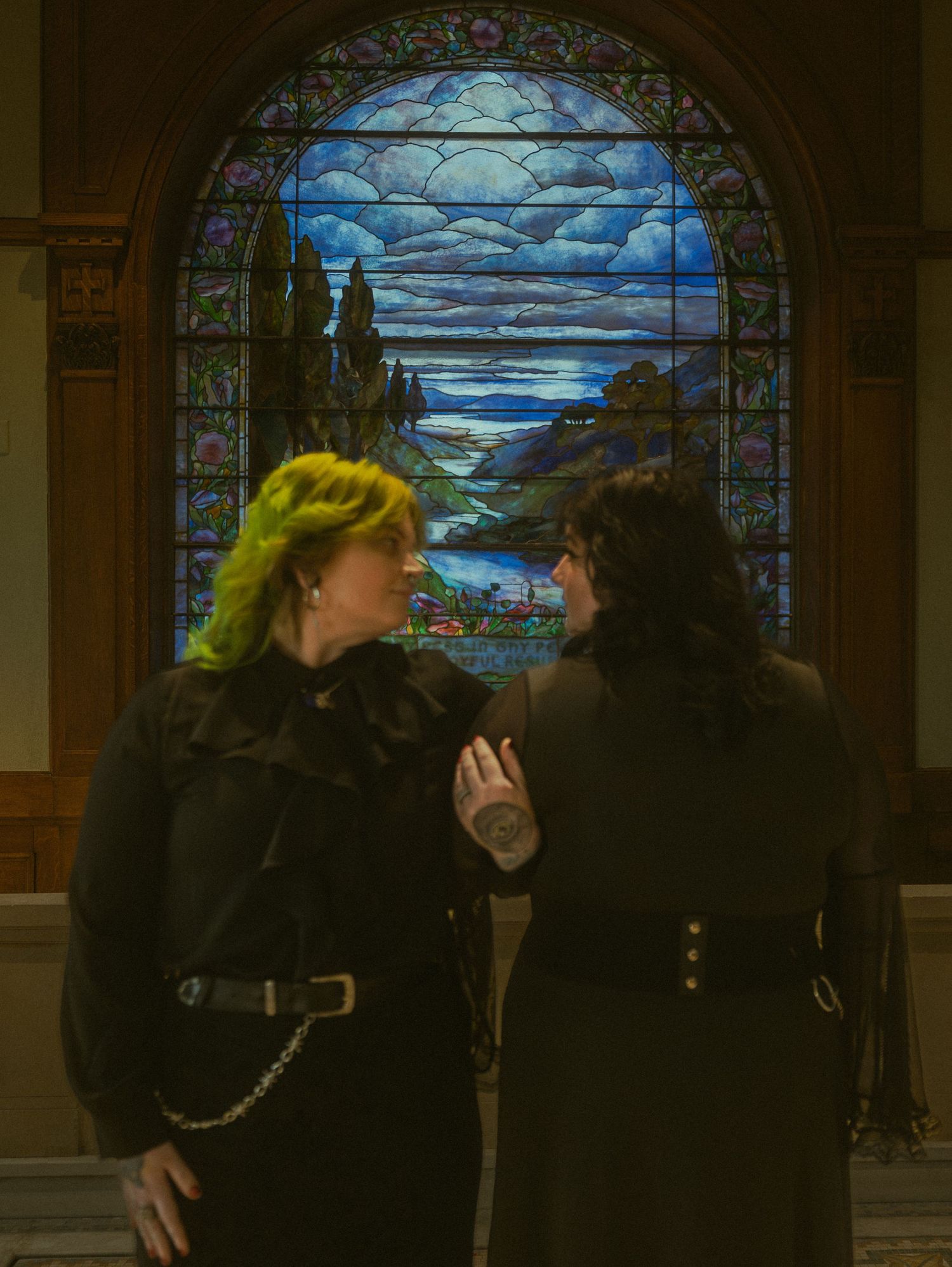 Two people in dark clothing stand before a beautiful stained glass window depicting a moonlit landscape.