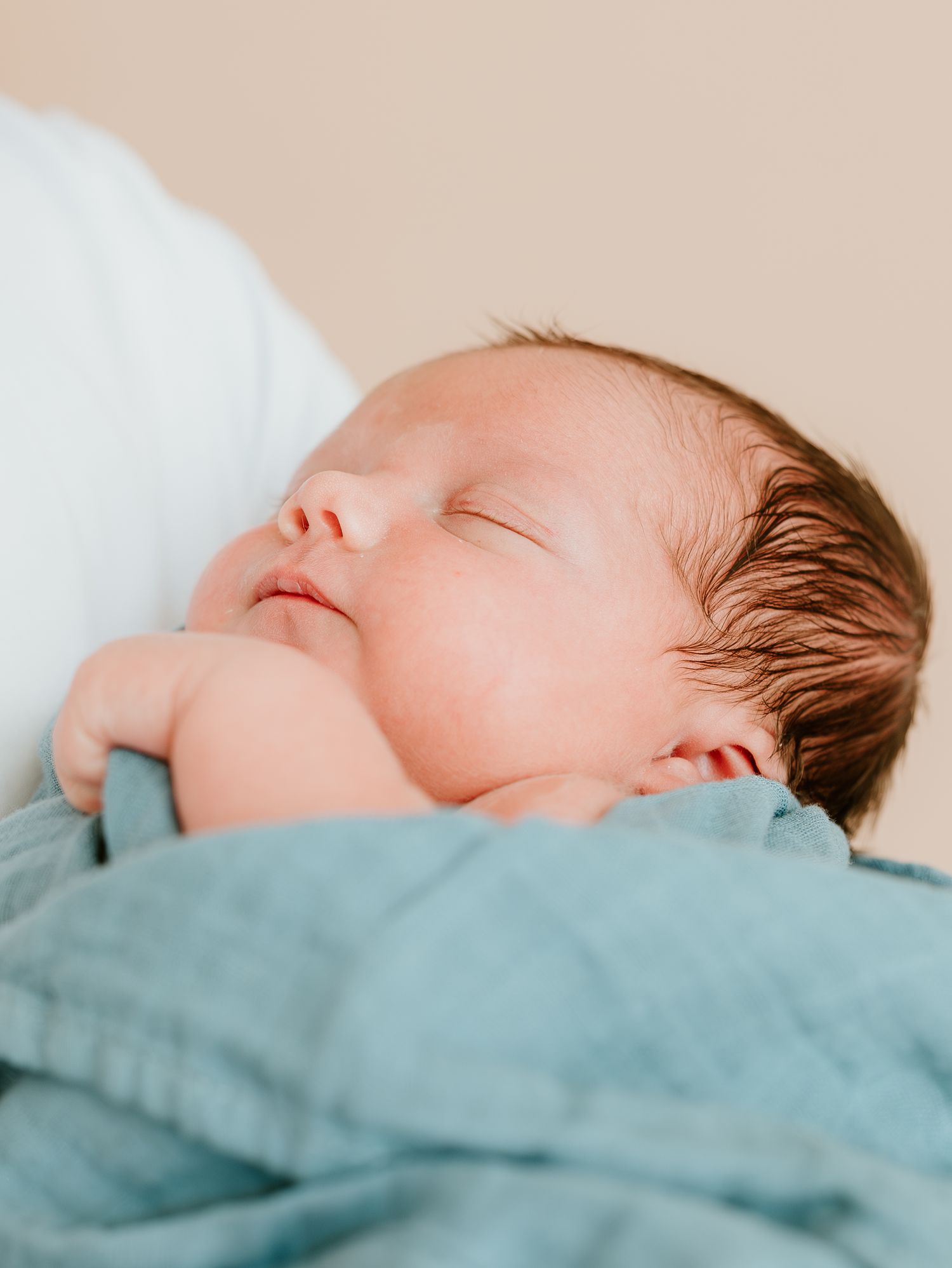 Sleeping newborn baby with brown hair rests peacefully on turquoise bedding in soft natural light.