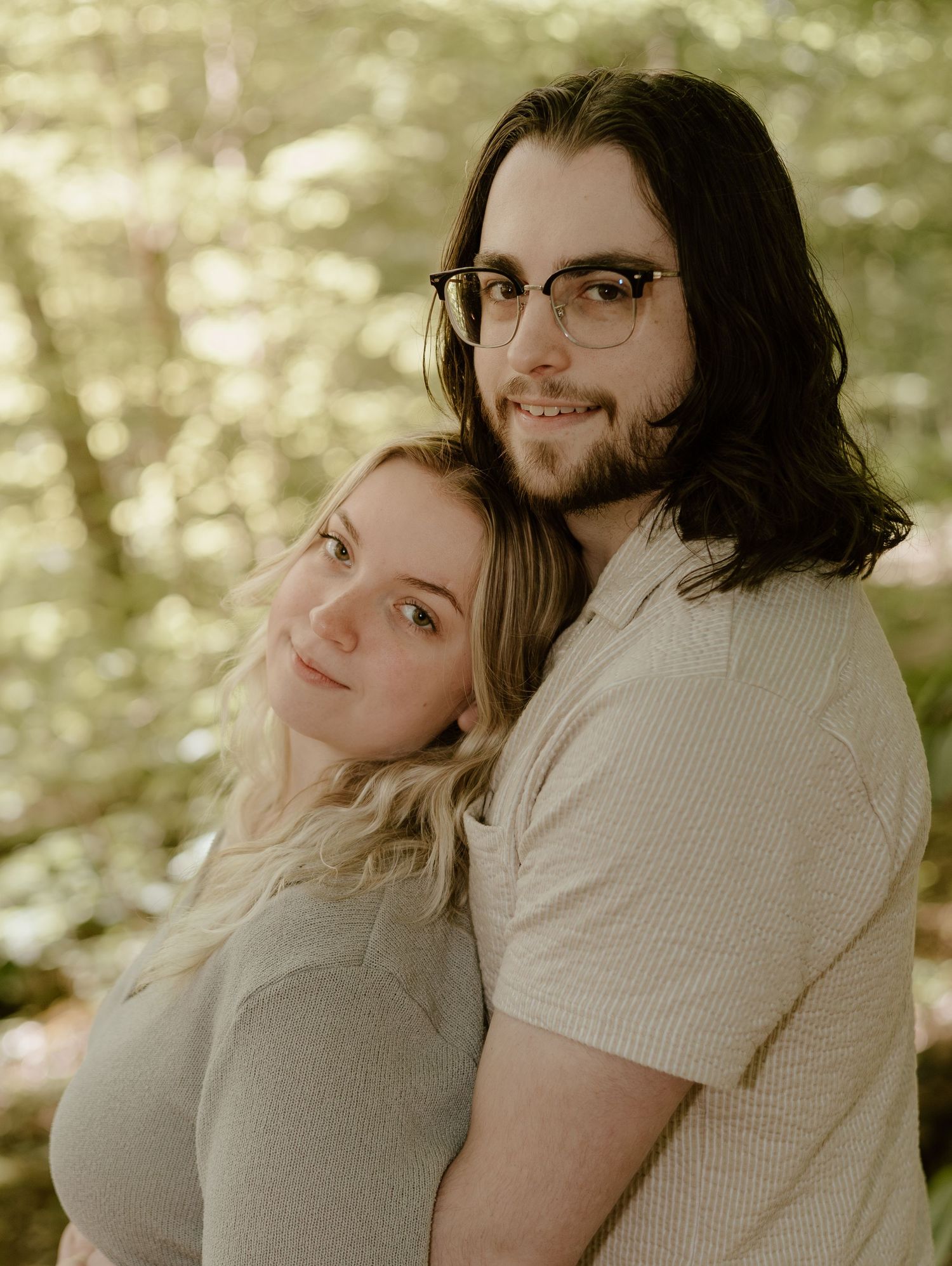 A romantic couple embraces in a sun-dappled forest setting with soft lighting.