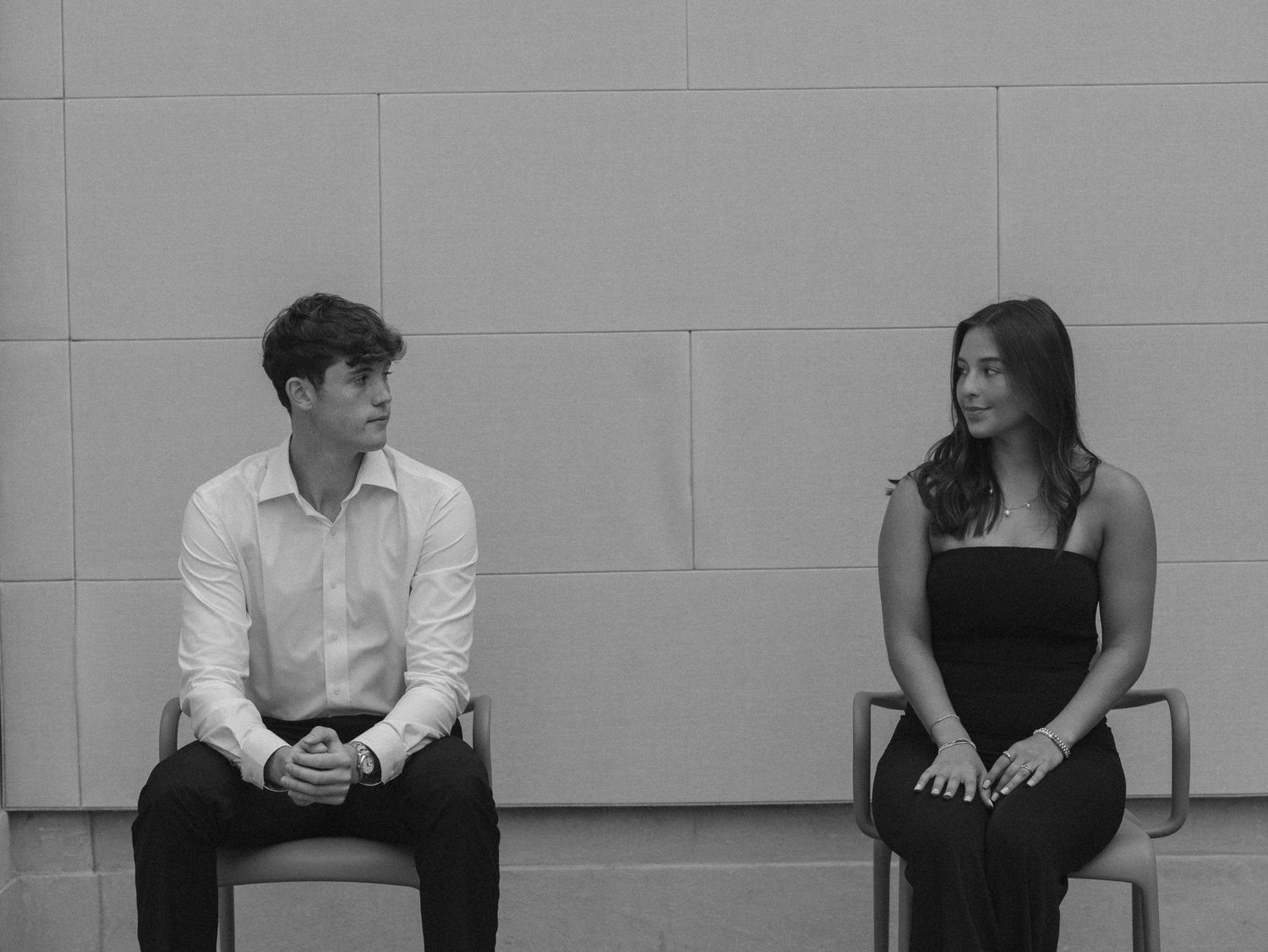 Two people sit on opposite sides of a white wall, creating a minimalist composition in black and white tones.