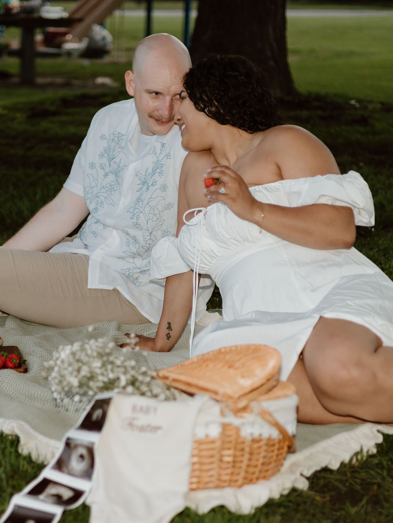 A romantic picnic scene on a blanket with a basket, flowers and ultrasound photos laid out in a grassy park.