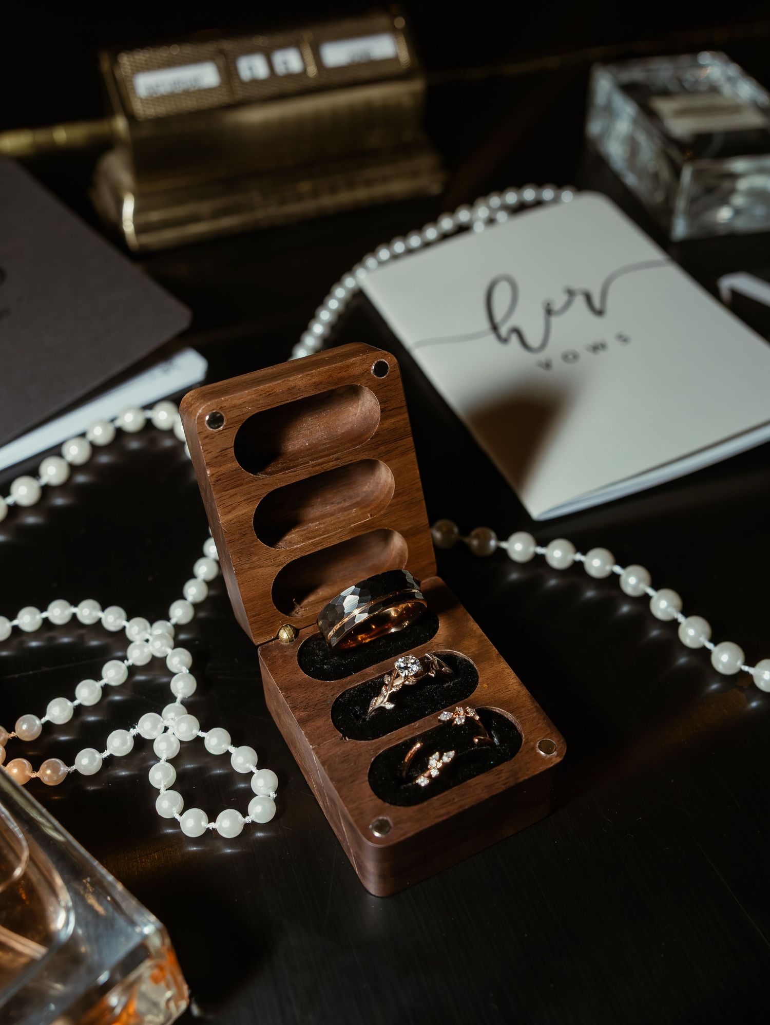 Elegant flat lay of wedding rings in wooden box with pearl necklace and her vows booklet.