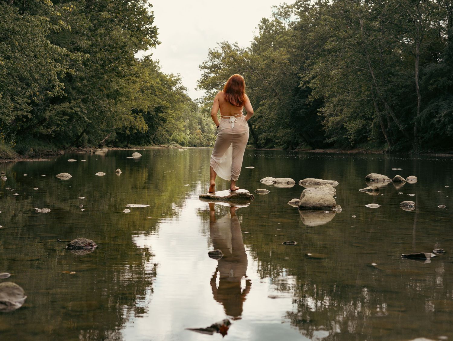 Person in a white dress walks through tranquil pond water surrounded by trees creating mirror-like reflections.