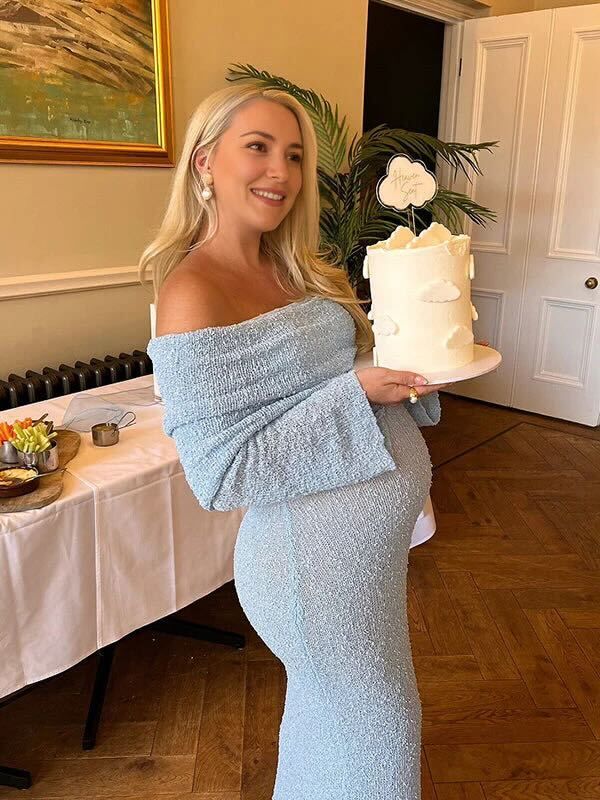 Person in a light blue off-shoulder dress holding a white tiered cake with floral decorations at an indoor event.