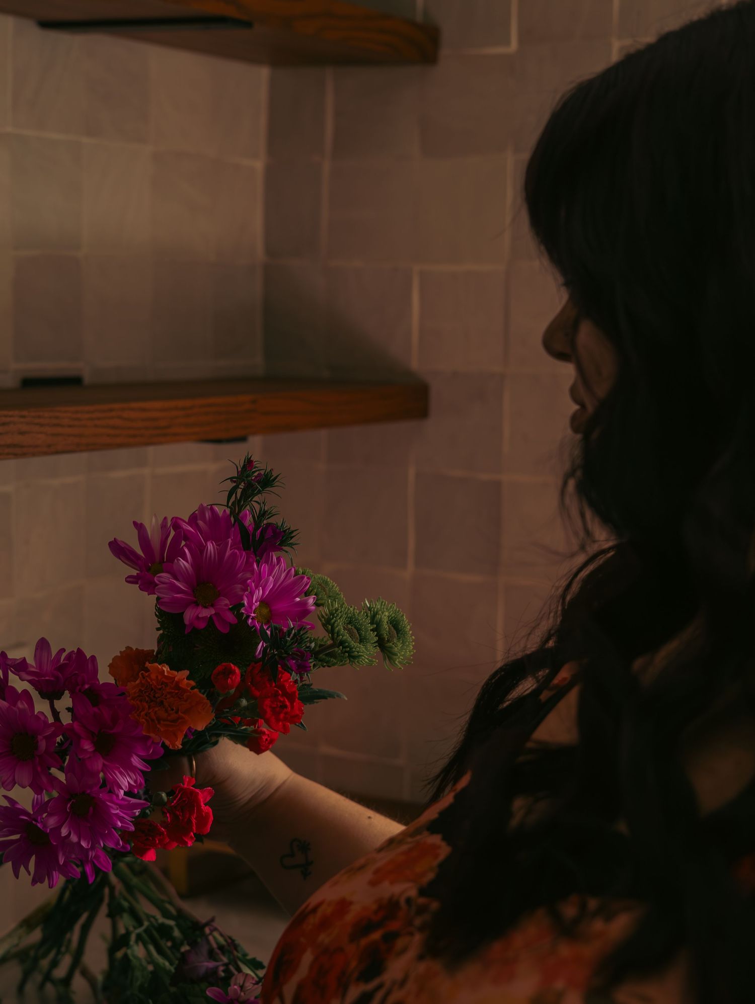 A person arranges vibrant purple and red flowers in a vase against a neutral wall with wooden shelves.