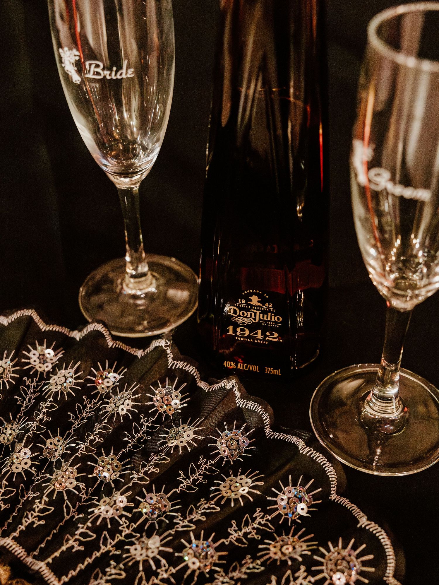 Elegant wedding setup featuring engraved bride and groom champagne flutes, Don Julio 1942 tequila, and a beaded black fan.
