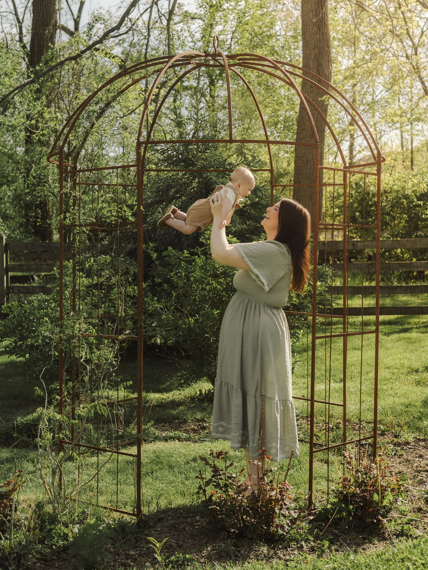 A mother in a sage green dress lifts her baby under a rustic garden arch surrounded by lush greenery.