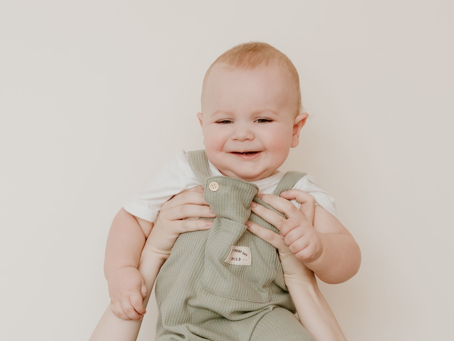 A happy baby wearing sage green overalls lays on a light beige background.