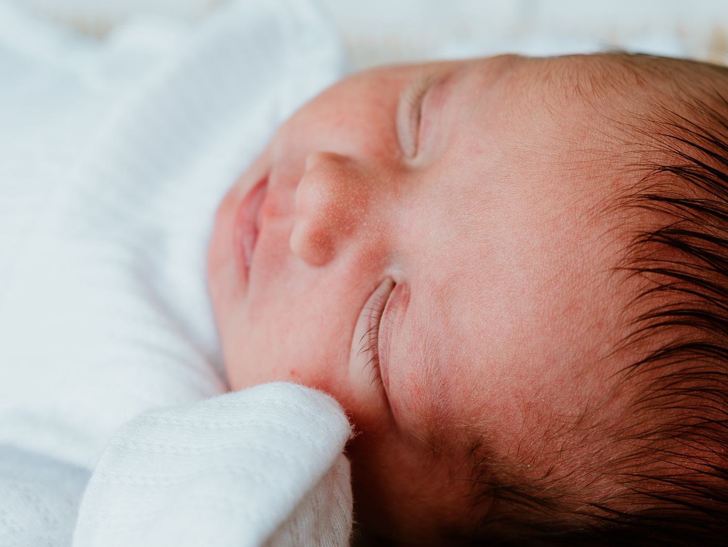 Close-up profile view of a sleeping newborn baby with dark hair against white bedding.