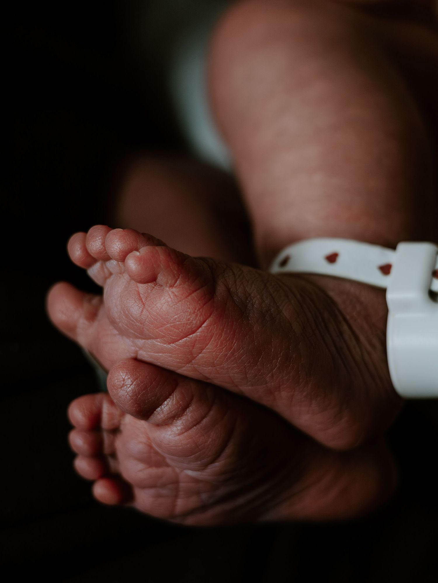 Close-up view of tiny newborn baby feet with white hospital ID bracelet against dark background.