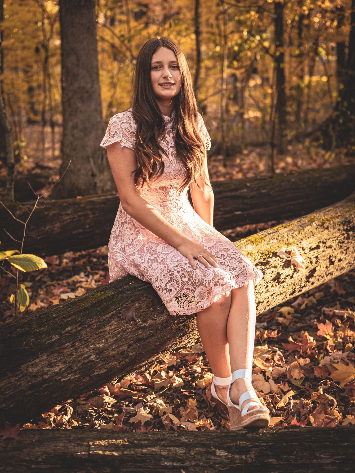 A person in a pink lace dress sits on a fallen log surrounded by autumn foliage in a golden-lit forest setting.