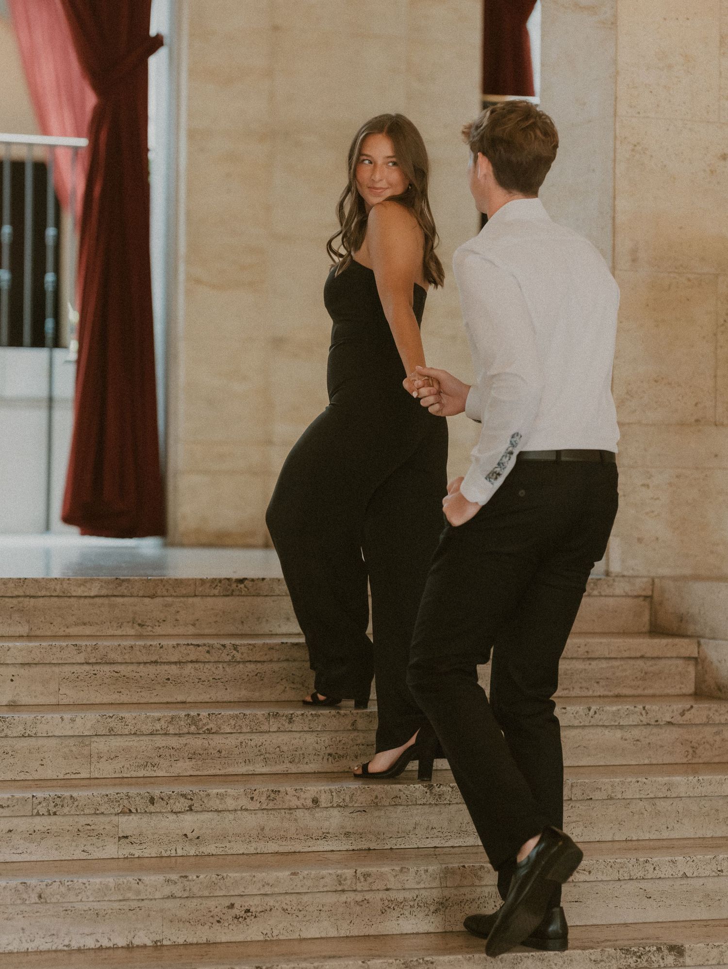 A couple in formal attire stands on marble steps having a conversation in an elegant indoor setting.