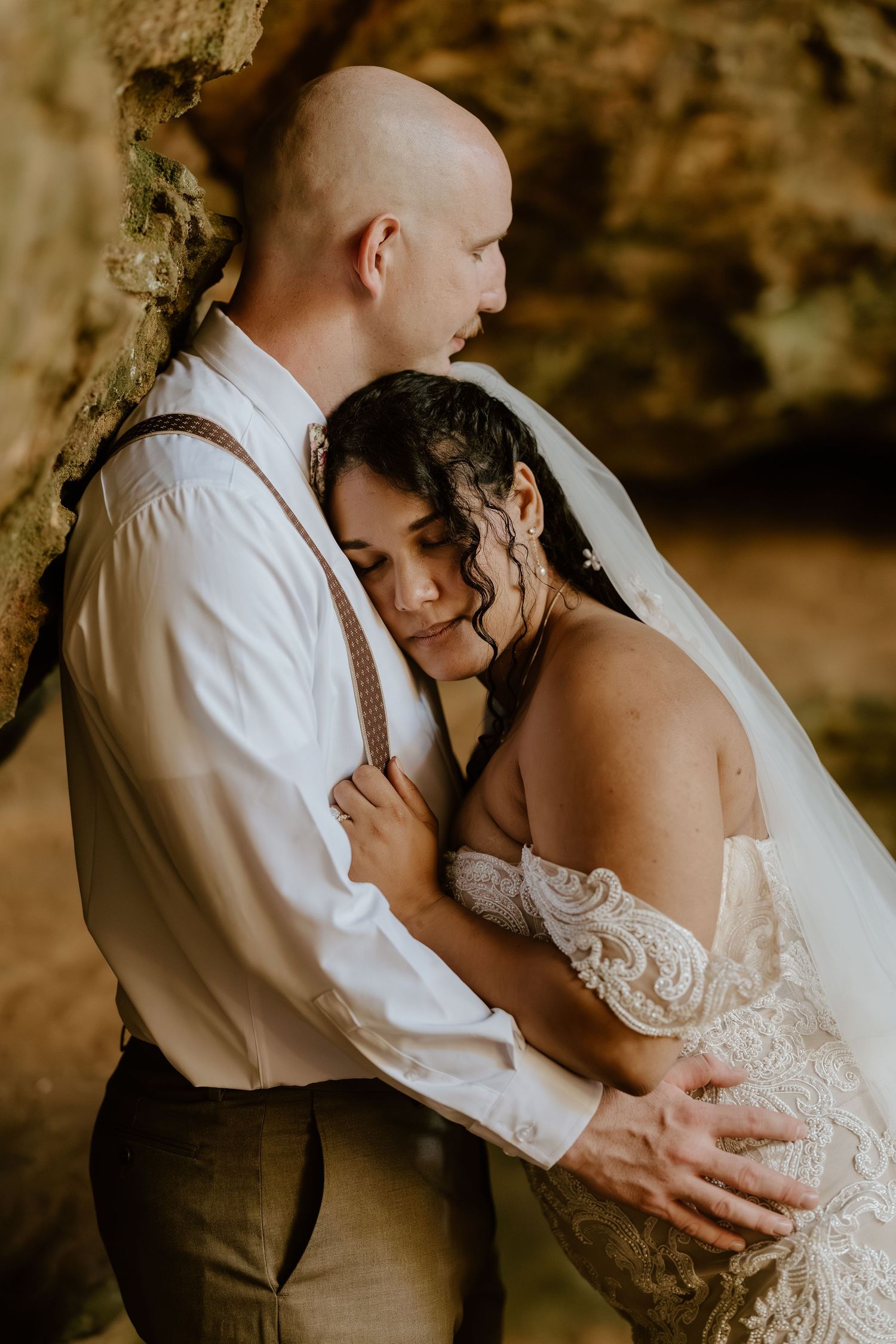 Intimate wedding portrait of couple embracing against textured stone wall in warm lighting.