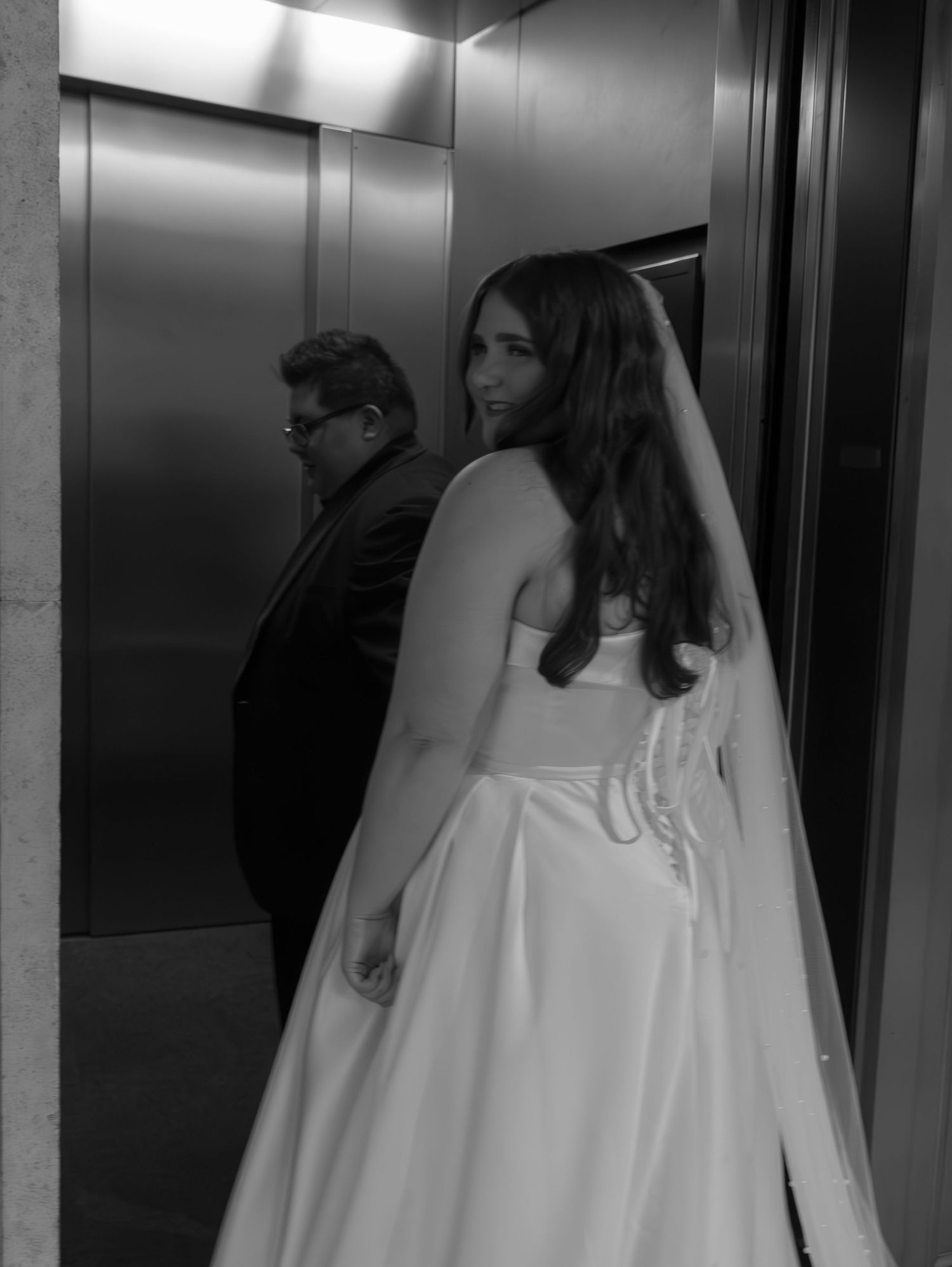 Bride in white wedding gown walking through modern elevator hallway.