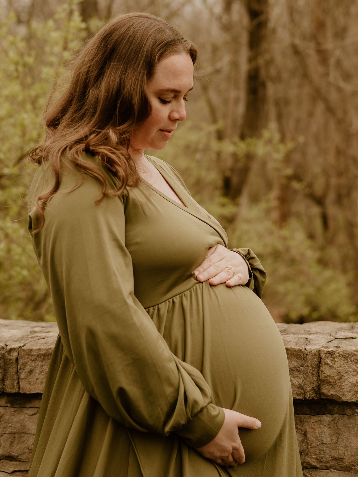 A pregnant woman in an olive green dress stands thoughtfully by a stone wall in an autumn forest setting.