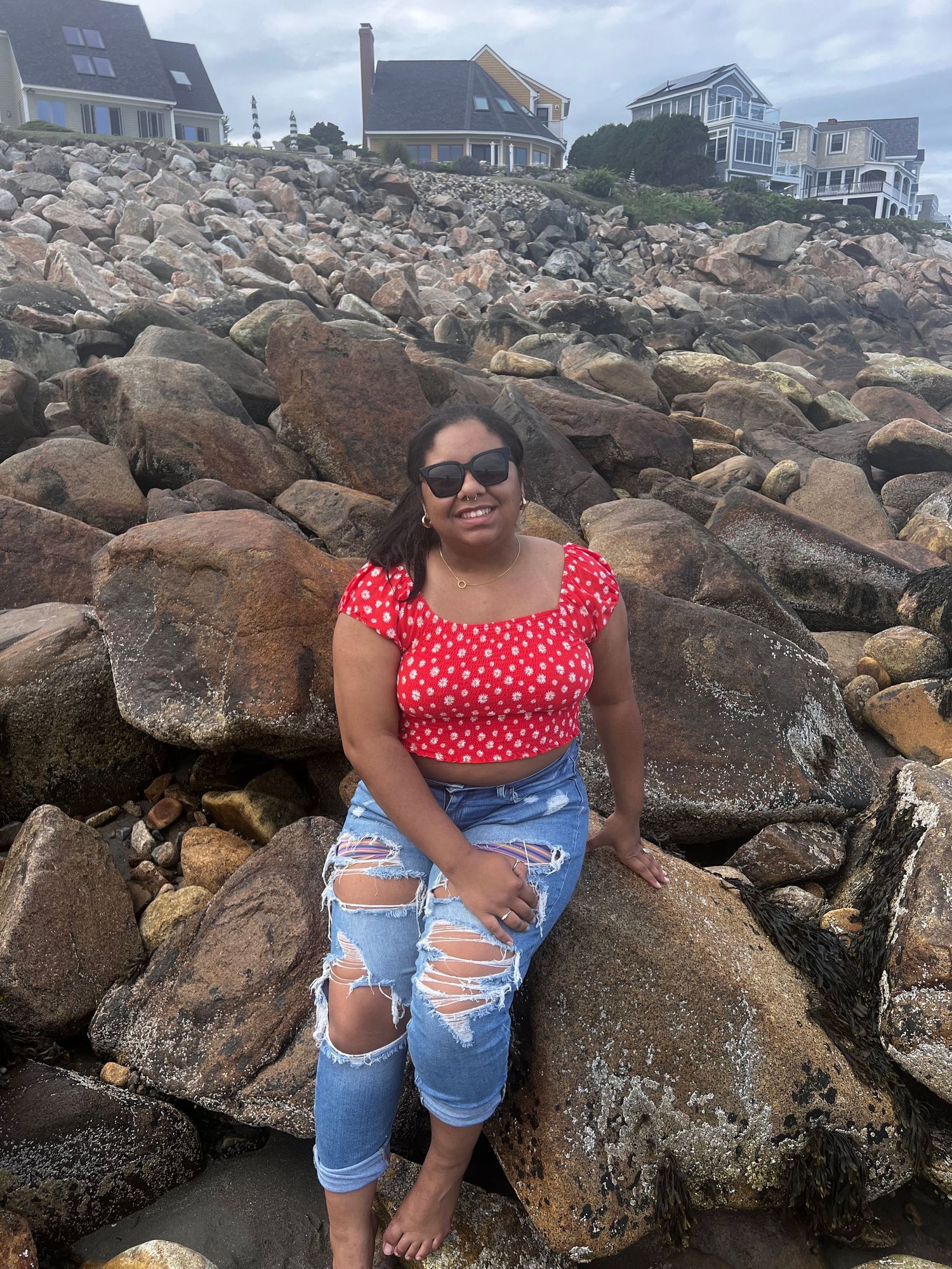 A person sitting on coastal rocks wearing a red polka dot top and ripped jeans near beachfront houses.