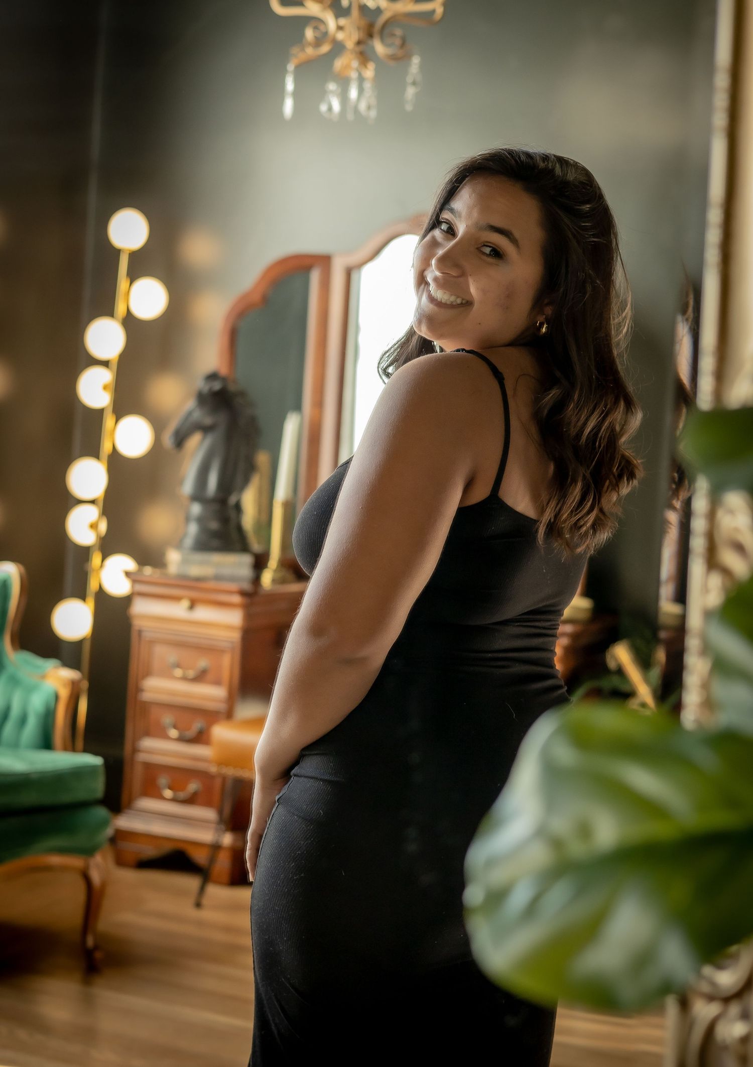 Woman in black dress smiling over shoulder in vintage room with mirror and warm string lights.
