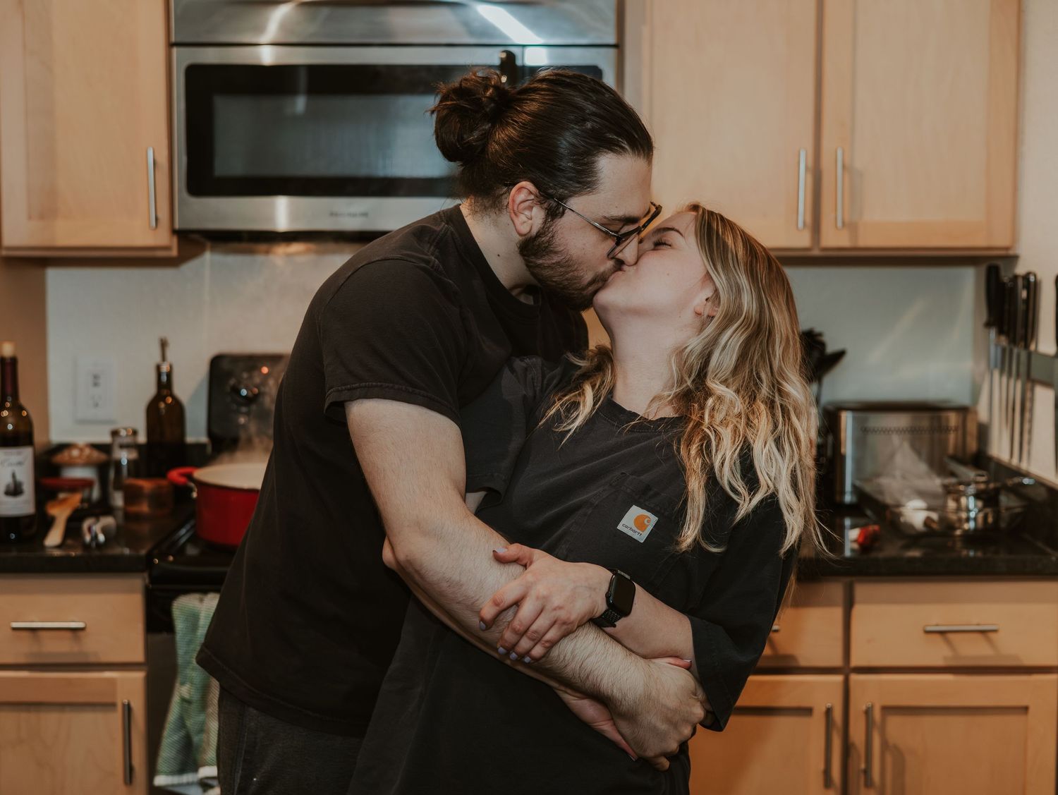 A couple shares an intimate kiss in a warmly lit kitchen setting.