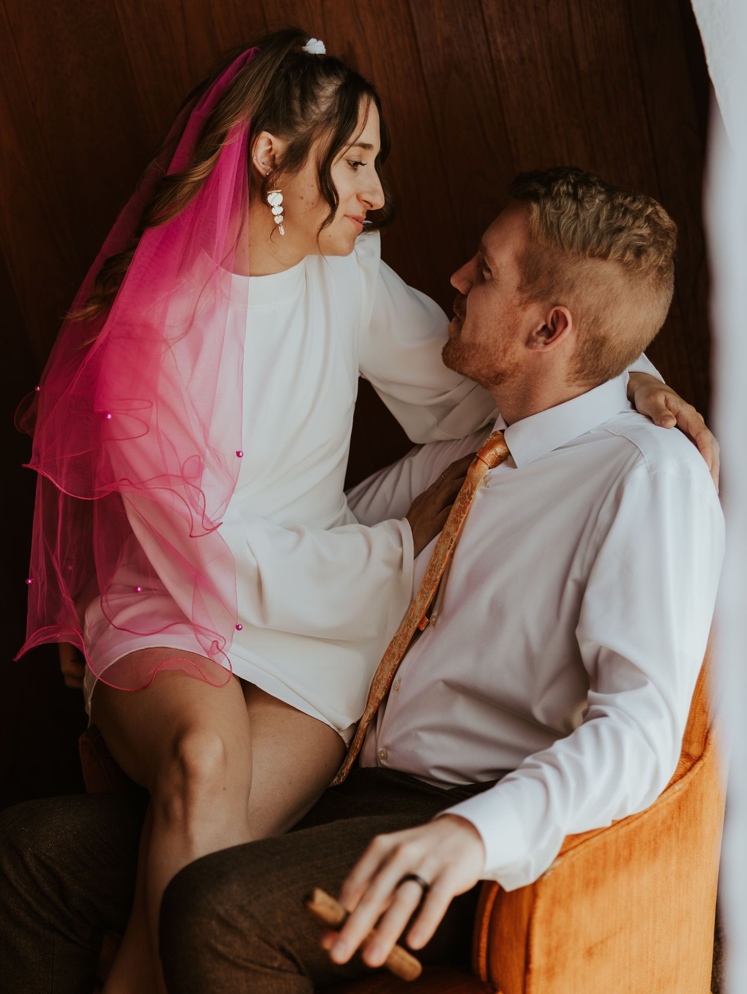 A couple in wedding attire embrace intimately during a romantic photoshoot with pink veil details.