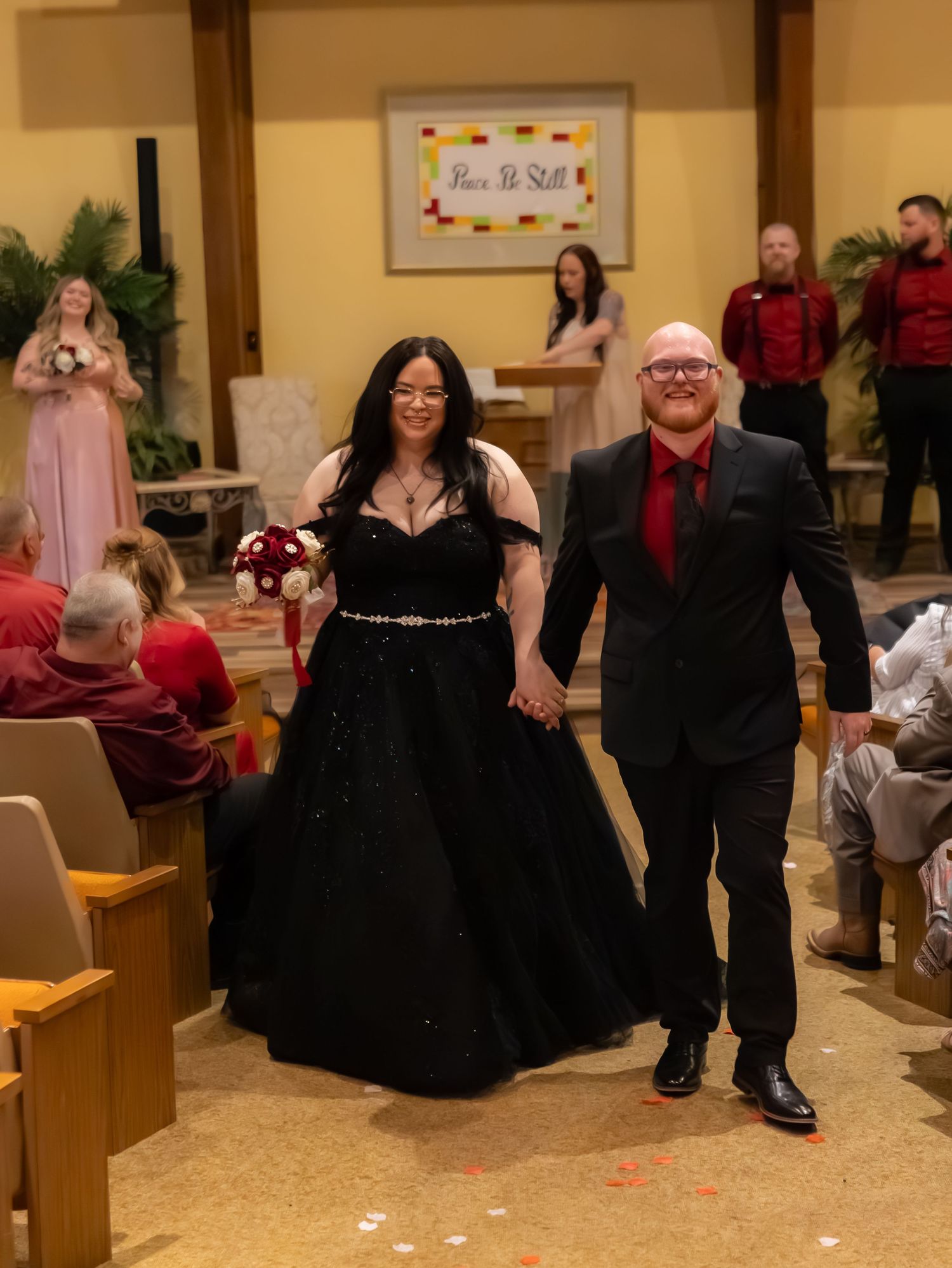 Newlyweds walk down the aisle, bride in black gown holding bouquet, groom in black suit with red tie.