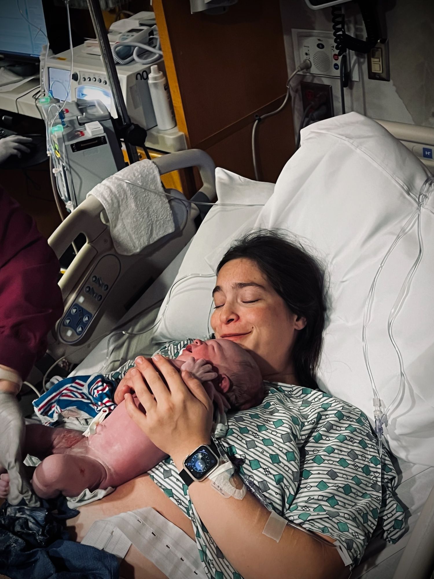 A newborn baby being held for the first time in a hospital room with medical equipment visible.