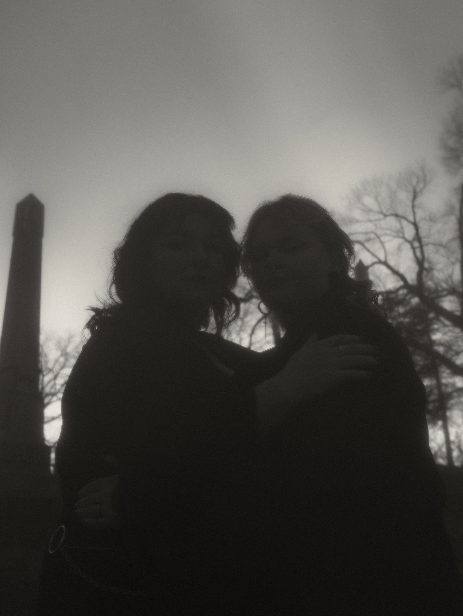 Silhouettes of cemetery monuments and trees against a misty grey sky at dusk.