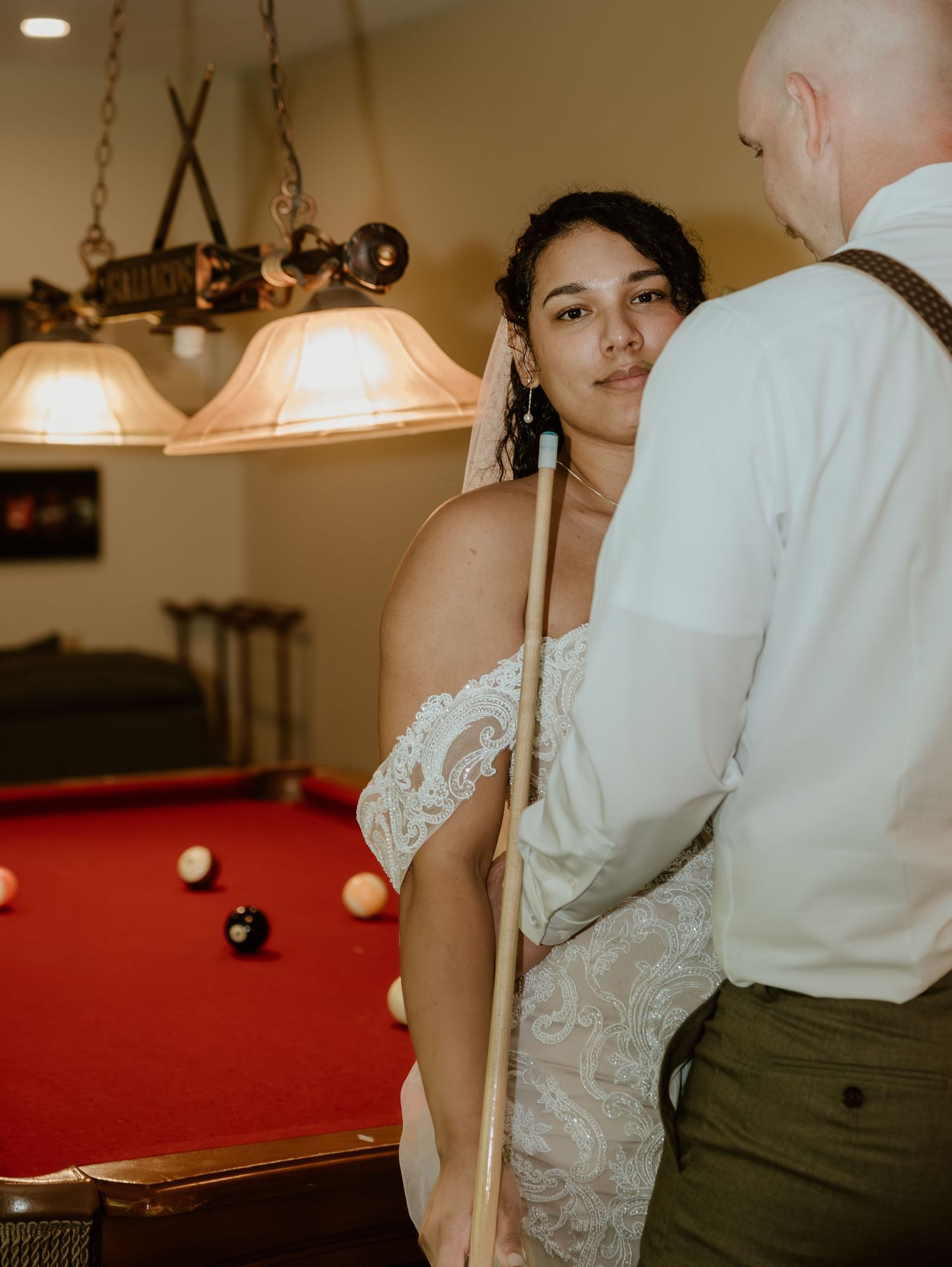Newlyweds in formal attire share an intimate moment near a pool table in warm indoor lighting.