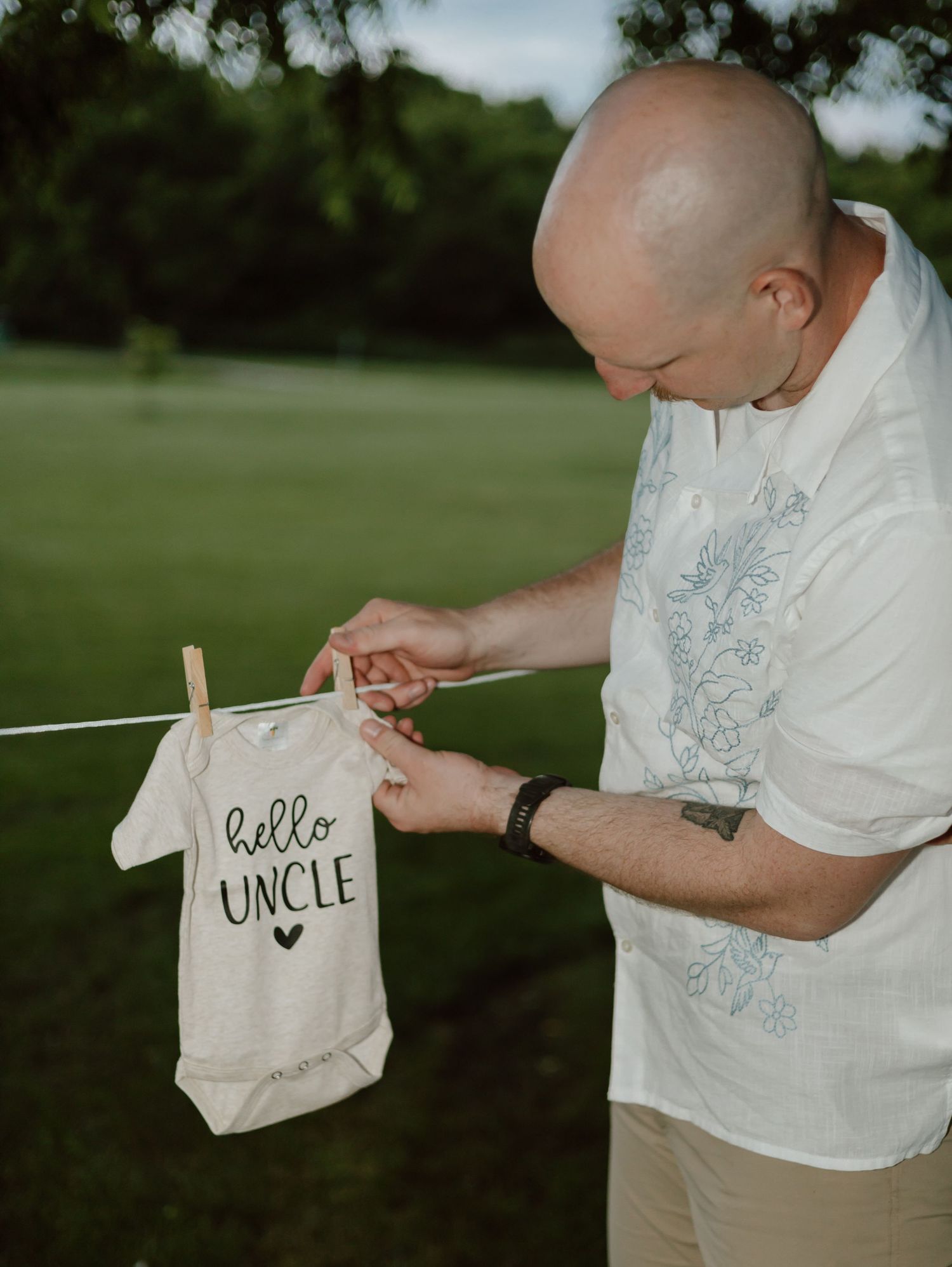 Someone pins a white baby onesie with 'Hello Uncle' text to a clothesline outdoors in a garden setting.