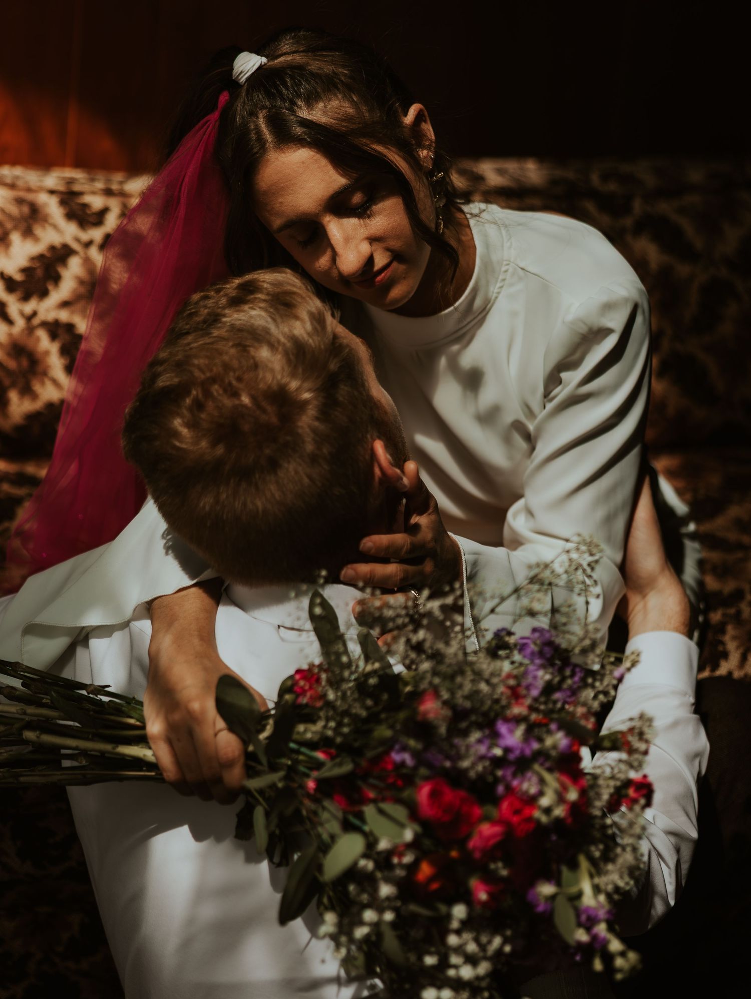 A romantic moment unfolds between a couple surrounded by deep red and purple florals in moody lighting.