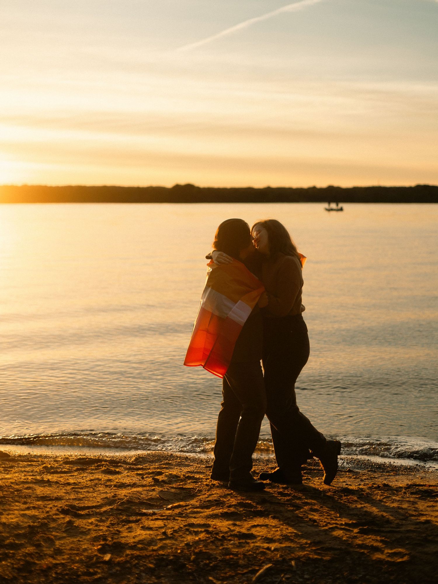 Two women wrapped in a blanket embrace on a sandy shore during a golden sunset over calm water.