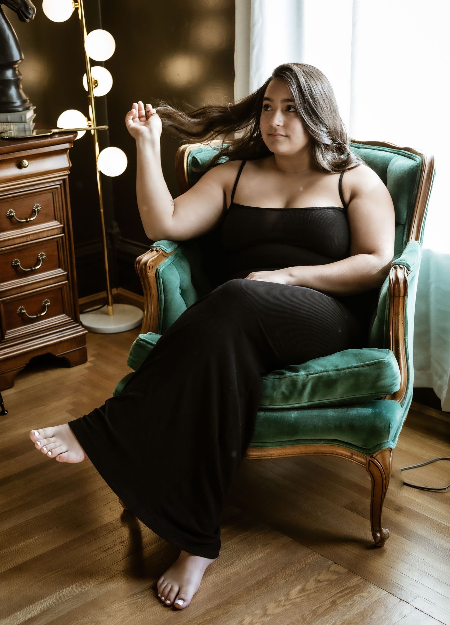 A woman in a black evening gown reclines in an emerald green vintage chair beside a dresser with string lights.
