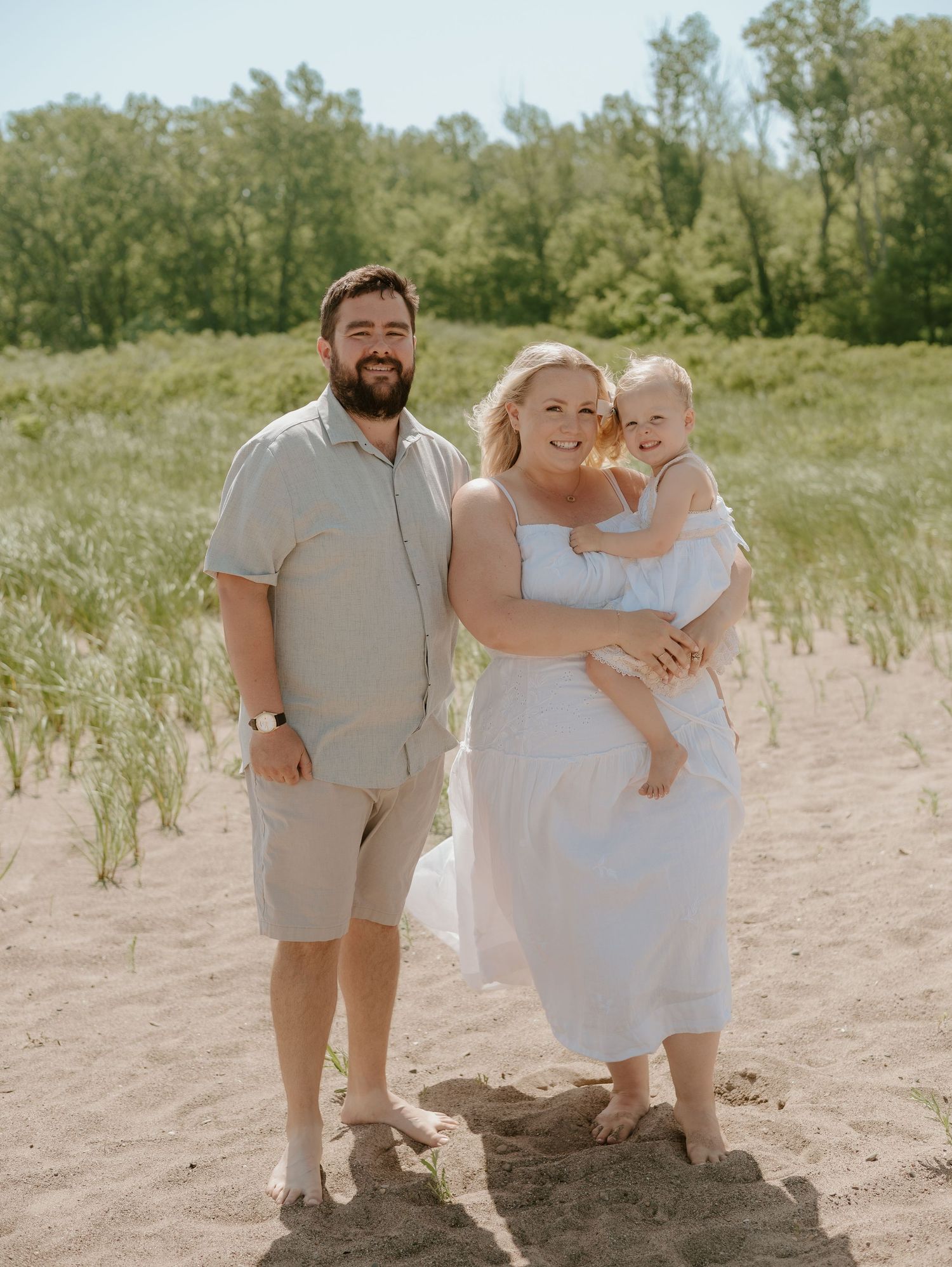 A happy family standing together on sandy beach dunes surrounded by green trees on a sunny summer day.