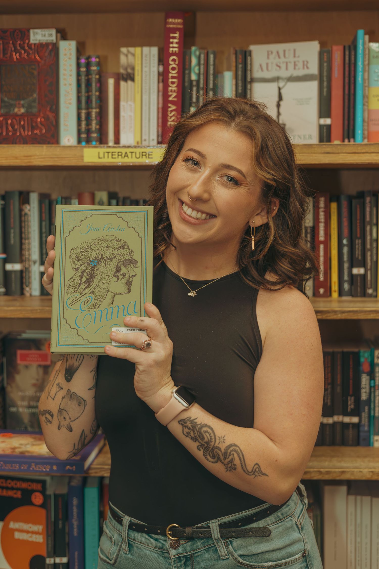 Person in black sleeveless top poses with books in front of library shelves showing off arm tattoos.