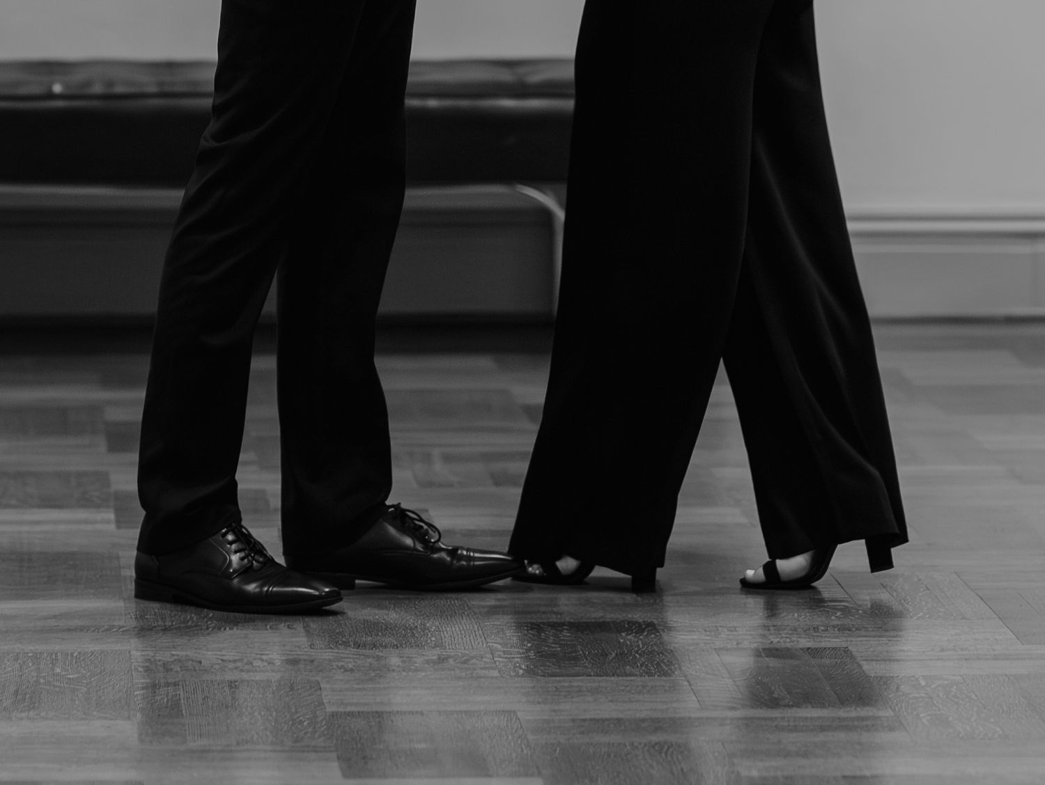 Dancers' feet in black formal shoes and pants glide across a polished dance floor during a ballroom performance.