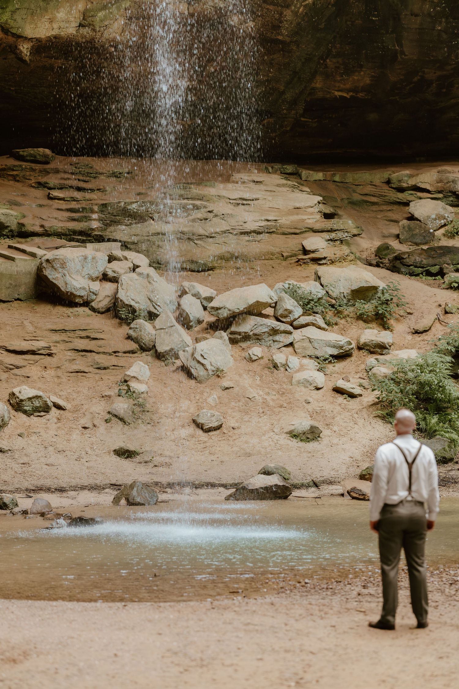 Wedding couple standing near a waterfall in a rocky natural setting.