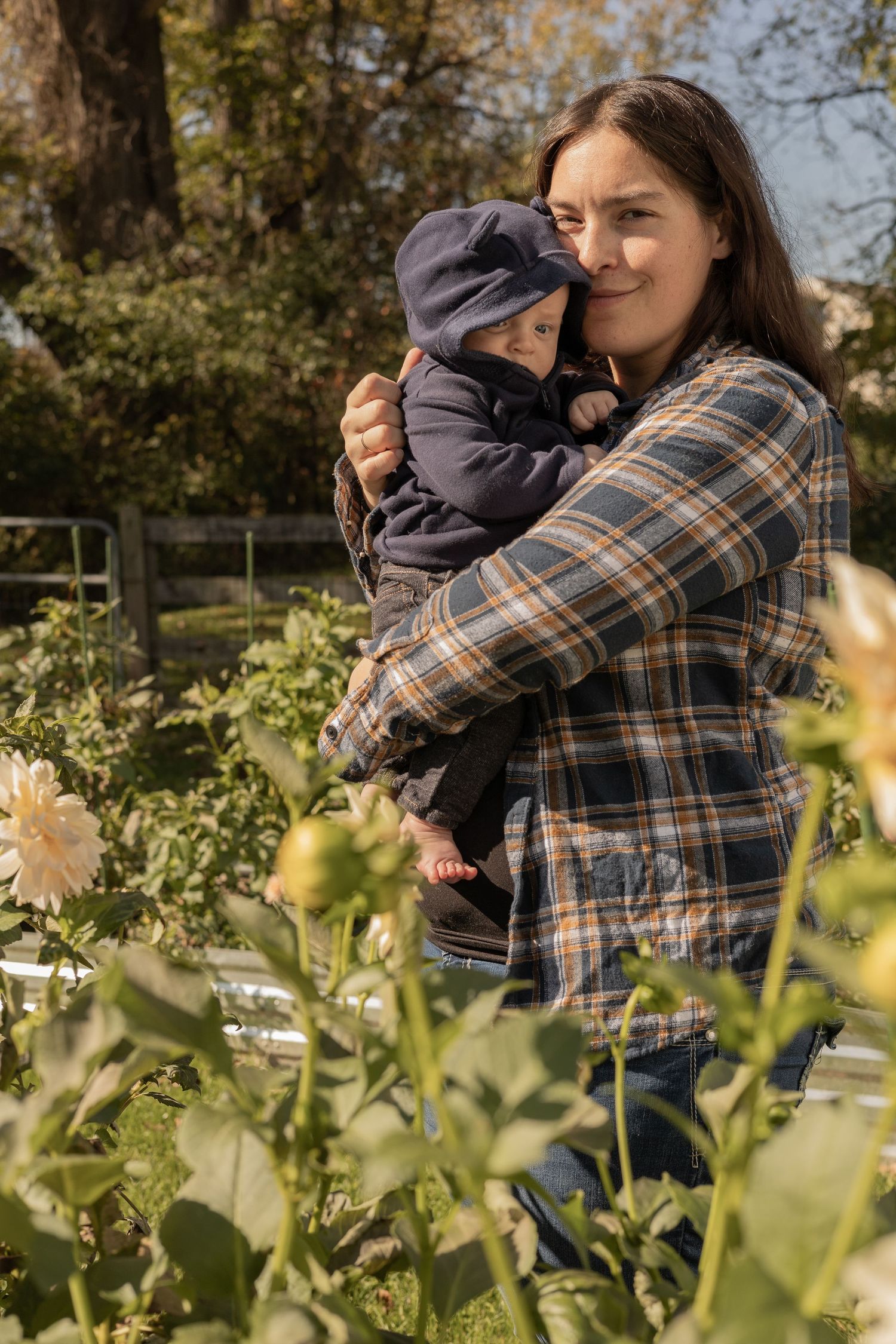 A person in a plaid flannel shirt holds a baby in a blue winter outfit while standing in a garden with flowers.