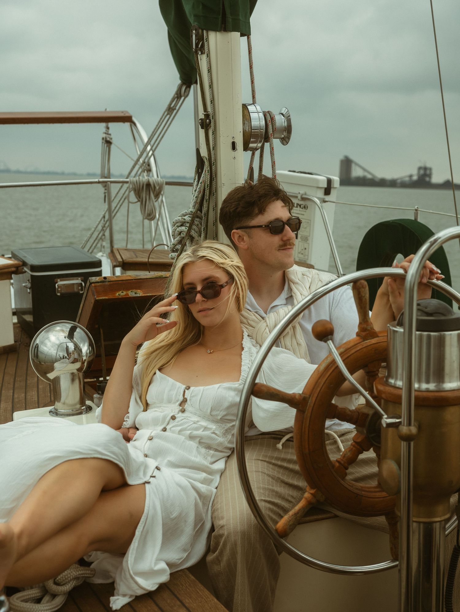 A couple in white outfits relaxes on a classic sailboat deck, woman in sundress leaning back as man steers the wooden helm.