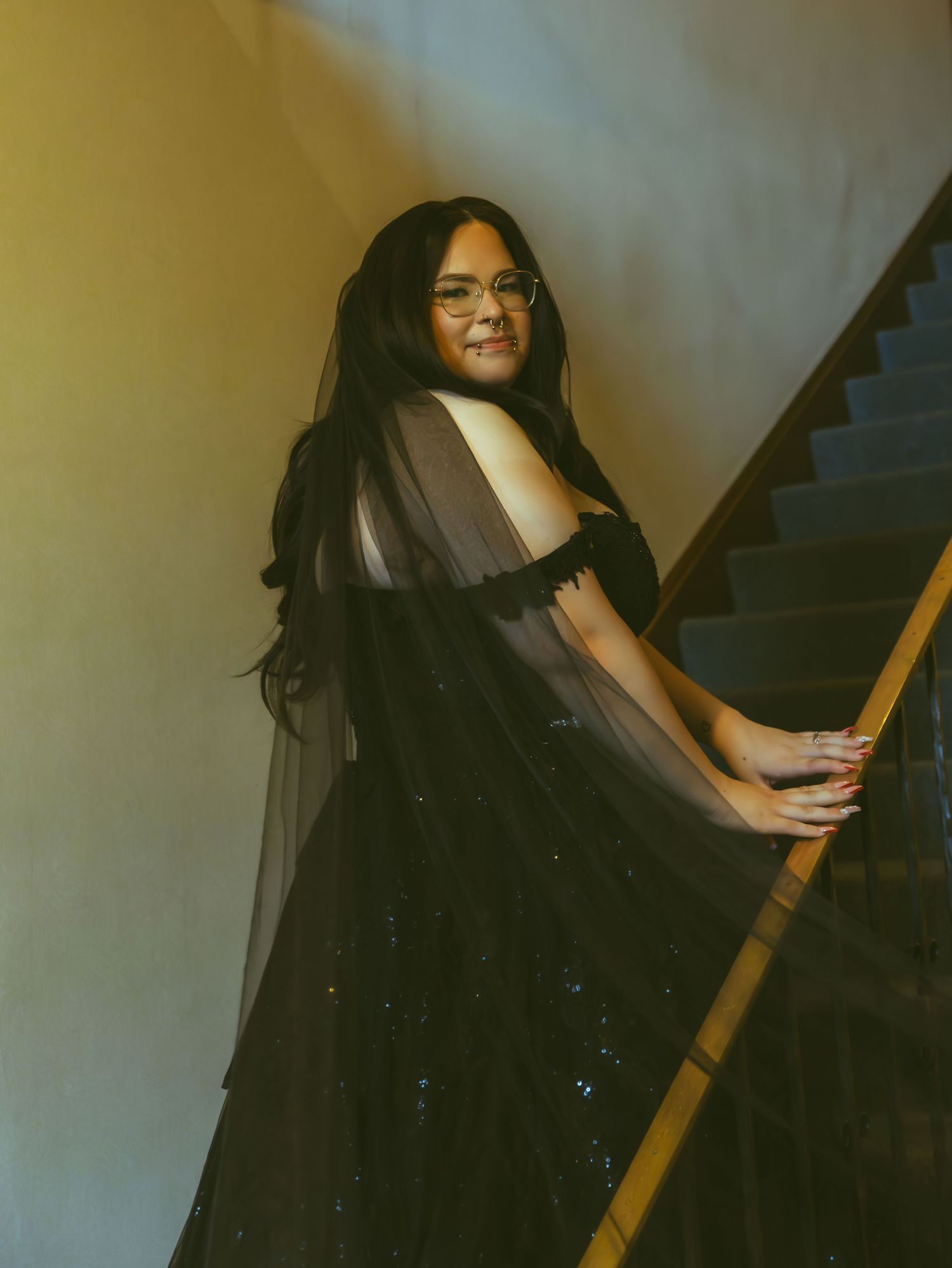 Bride poses elegantly on staircase in stunning black tulle off-shoulder wedding gown with veil.