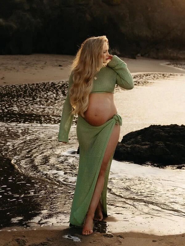 Silhouetted against a beach sunset, an expectant mother in a sage green dress poses gracefully near the water's edge.