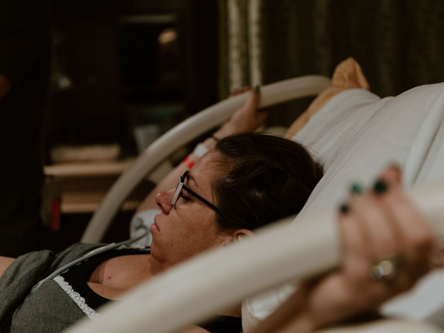 A woman wearing glasses rests in a hospital bed, gripping a bed rail during labor.
