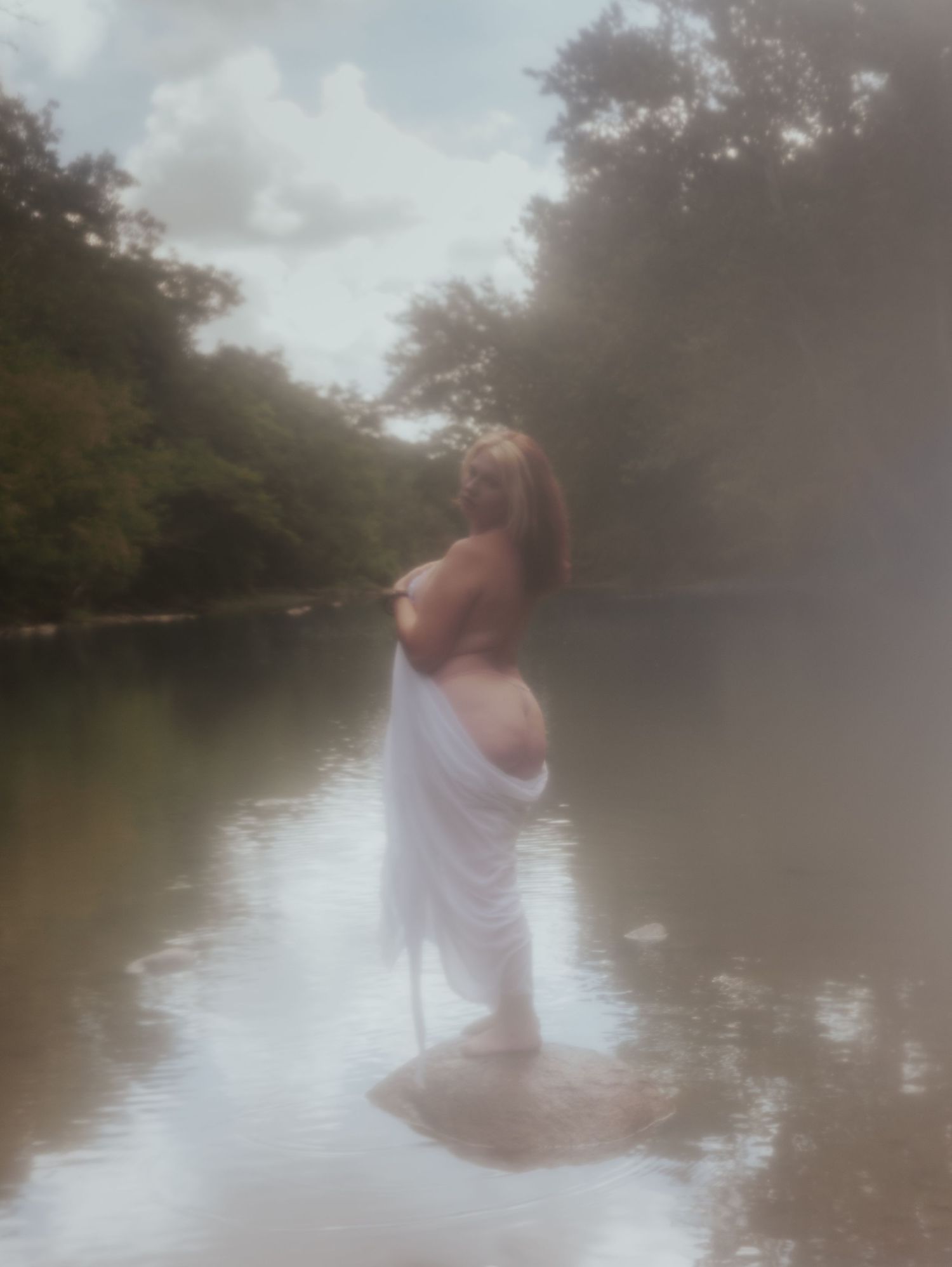 A figure draped in white fabric stands in misty water surrounded by trees creating an ethereal nature scene.