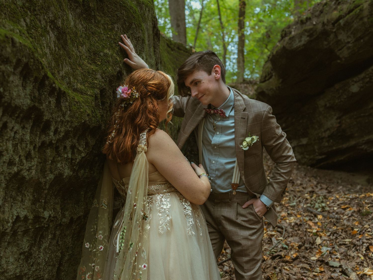 A romantic woodland wedding scene with a couple posing between mossy rock formations.