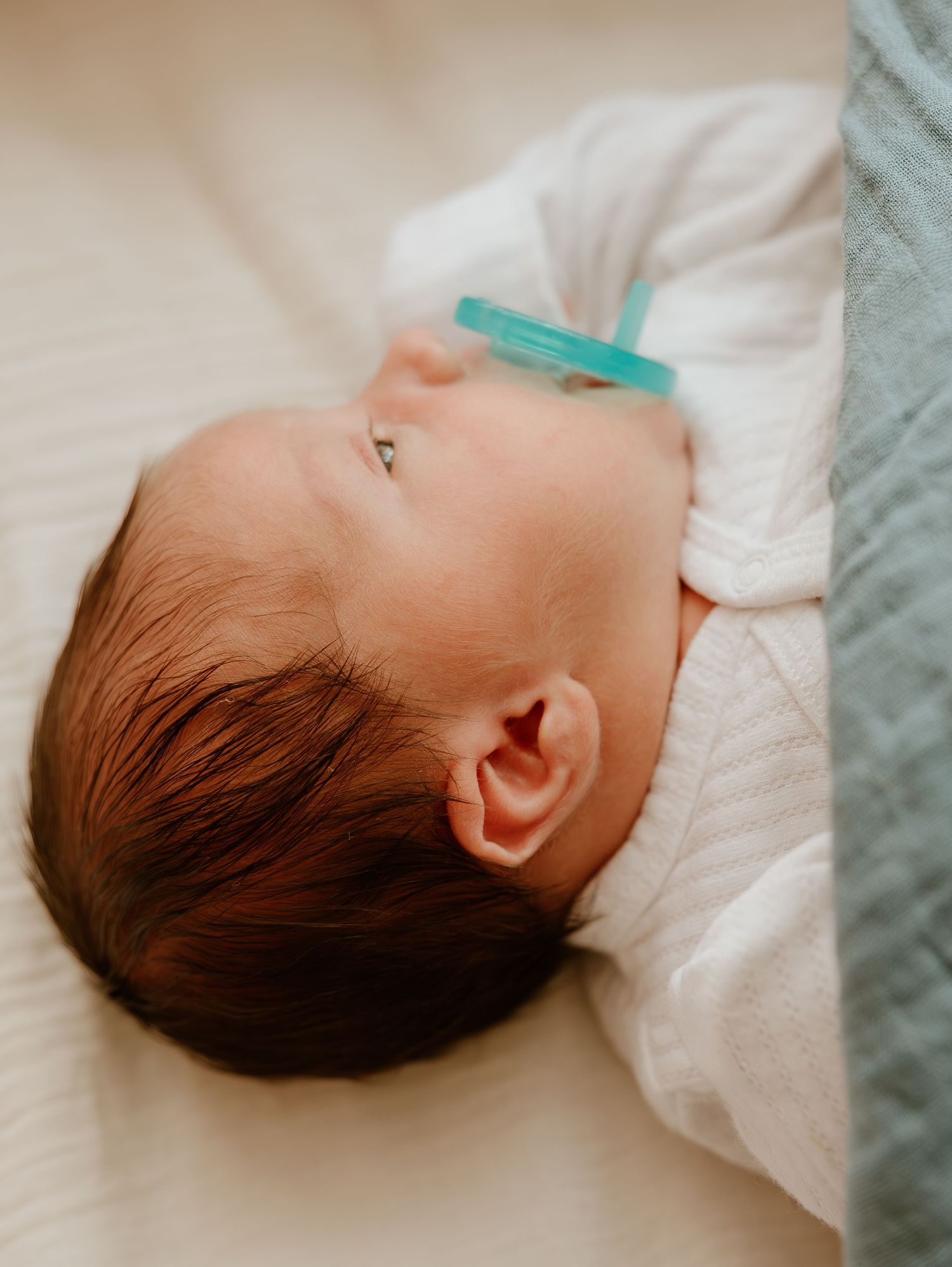 A sleeping infant with dark hair has a turquoise pacifier while resting on white bedding.