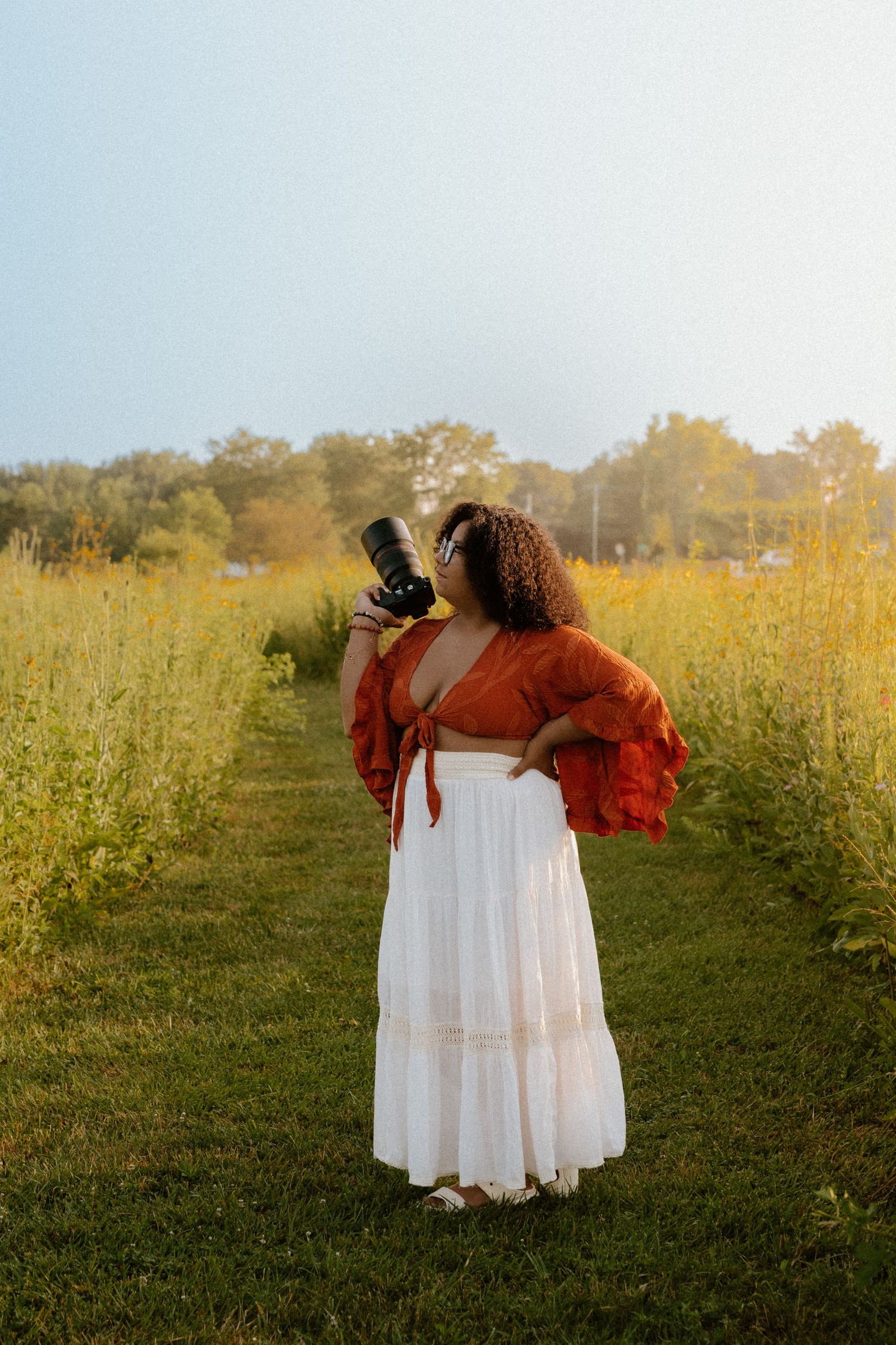 Two individuals in flowy white dresses standing together in a golden summer field at sunset.