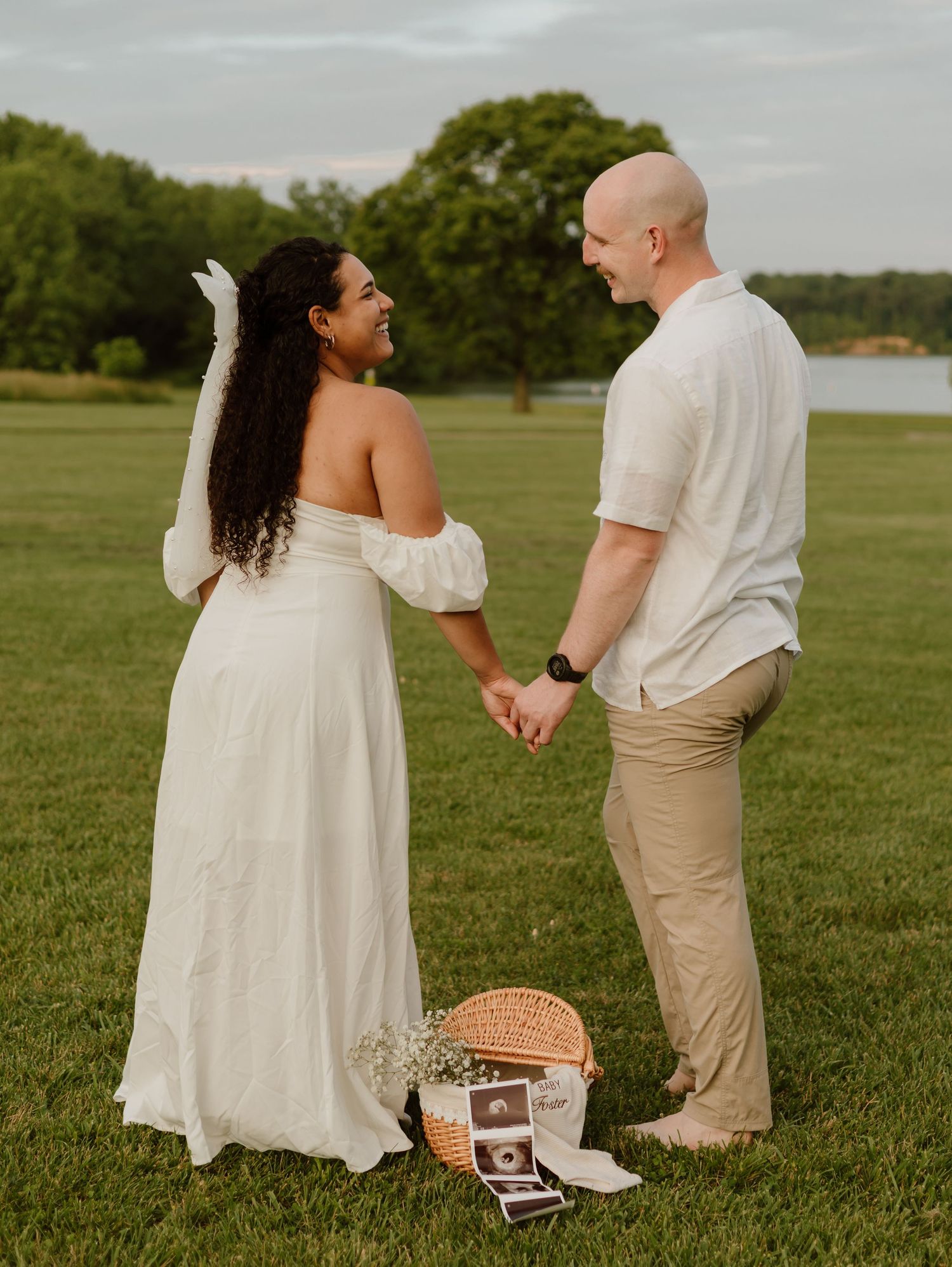 A couple in white and khaki outfits hold hands while standing in a grassy field during golden hour.