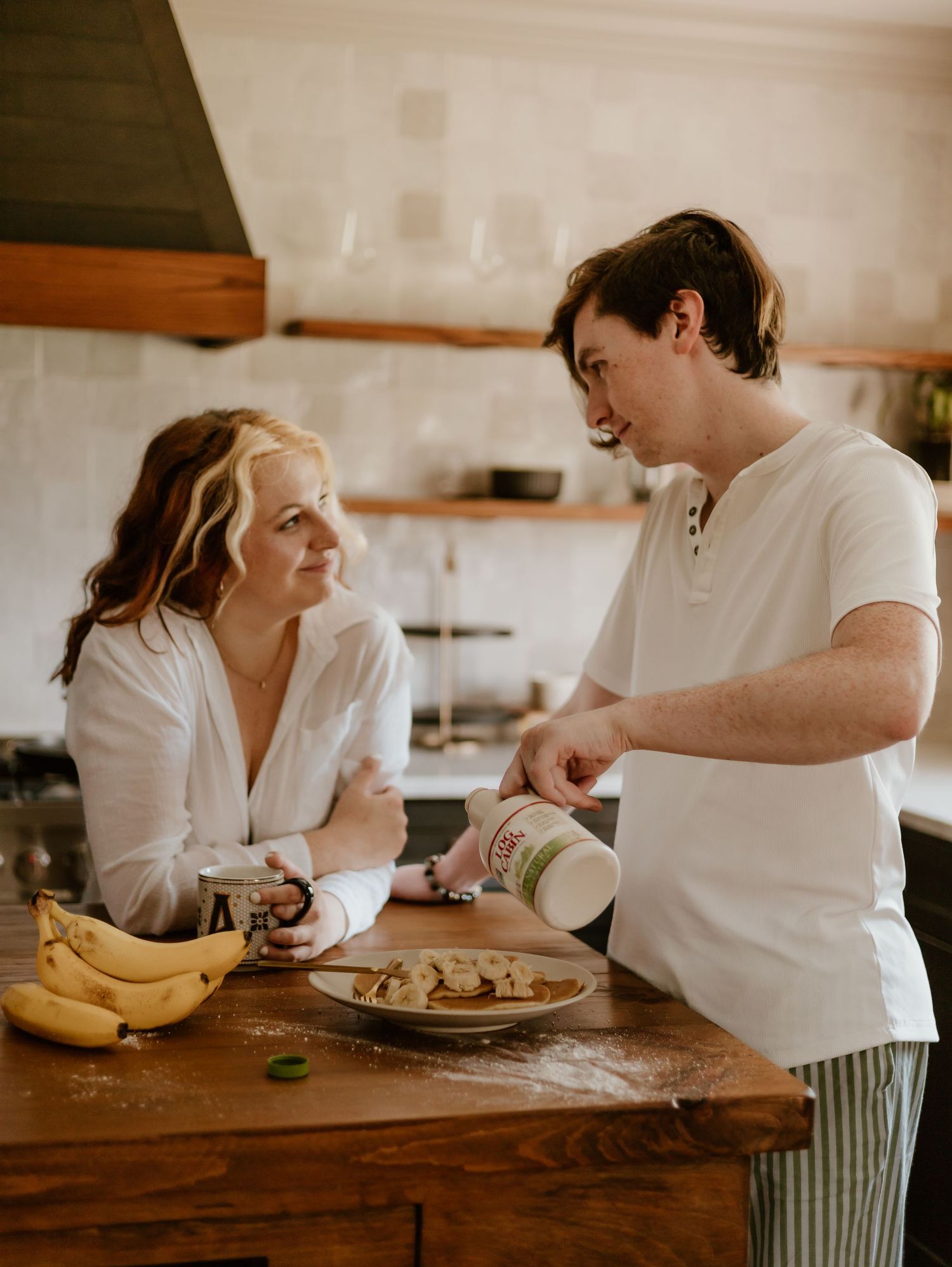 Two people having a casual conversation while preparing food at a rustic wooden kitchen counter with bananas nearby.